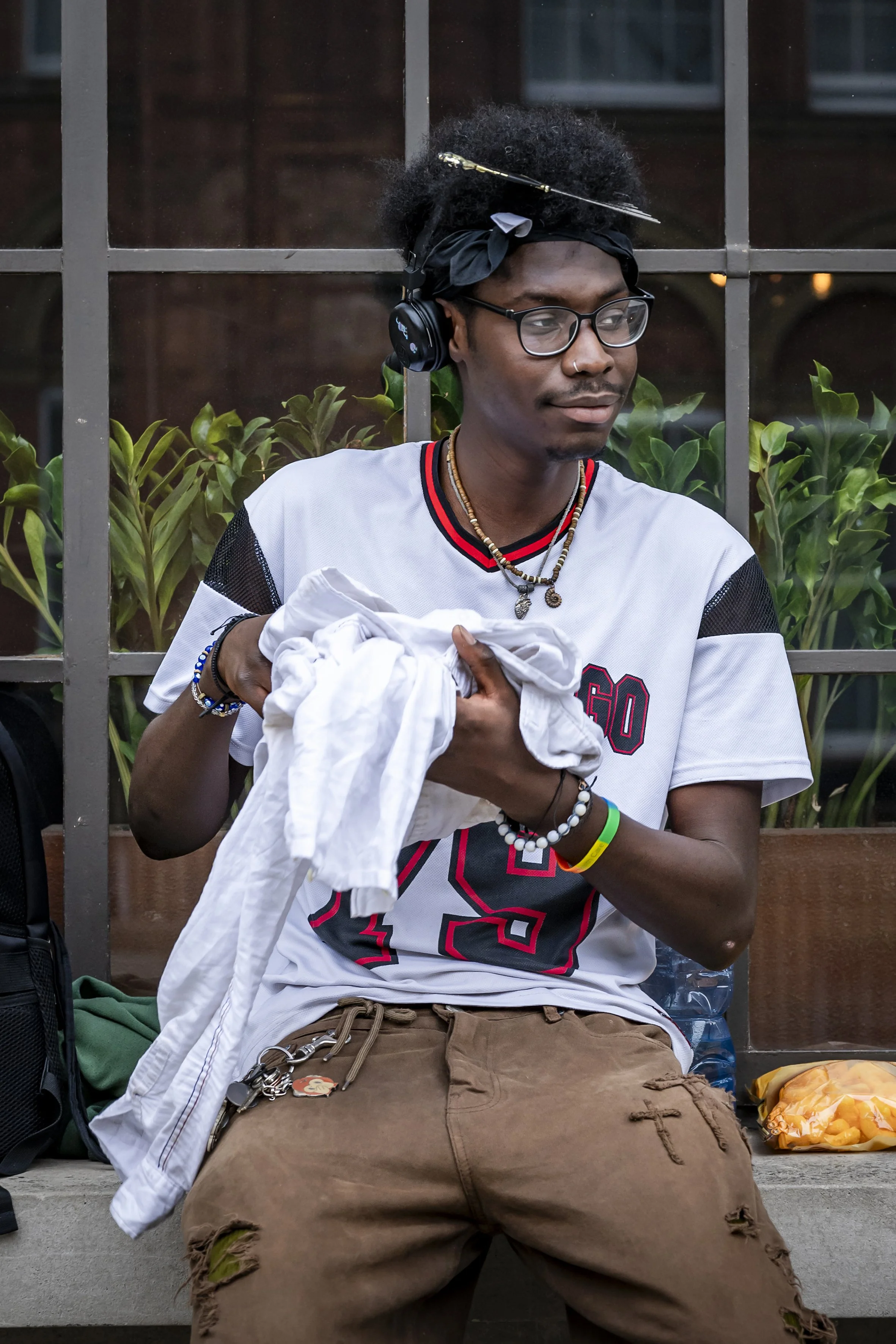 A young man with glasses, wearing headphones, a white sports jersey, and brown ripped pants, sitting on a bench, holding a white cloth or clothing item, with a background of green plants and a window with multiple panes.