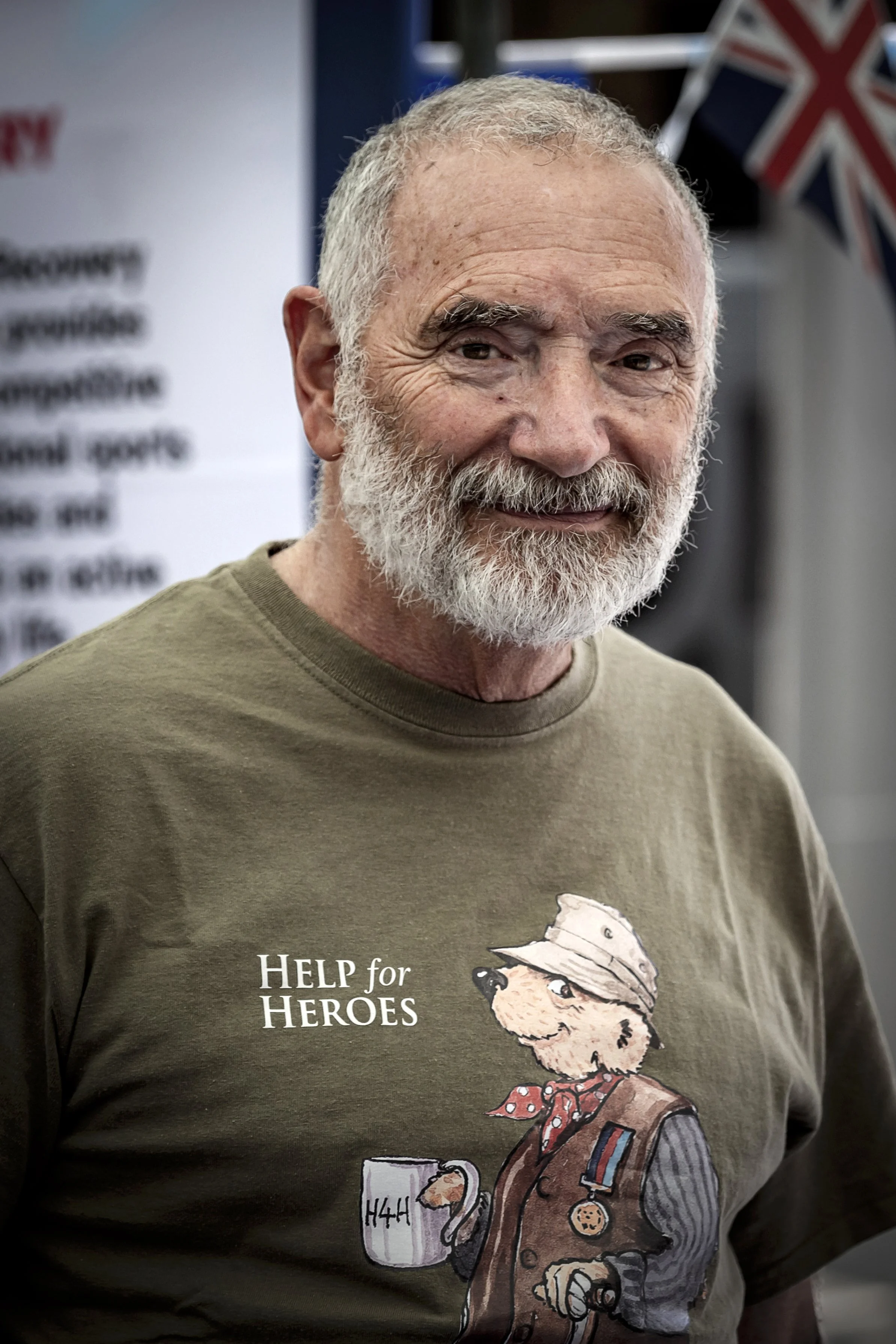 An older man with gray hair and a beard smiling, wearing a brown T-shirt with a cartoon dog dressed as a veteran holding a mug with 'H4H' on it, and the text 'Help for Heroes'.