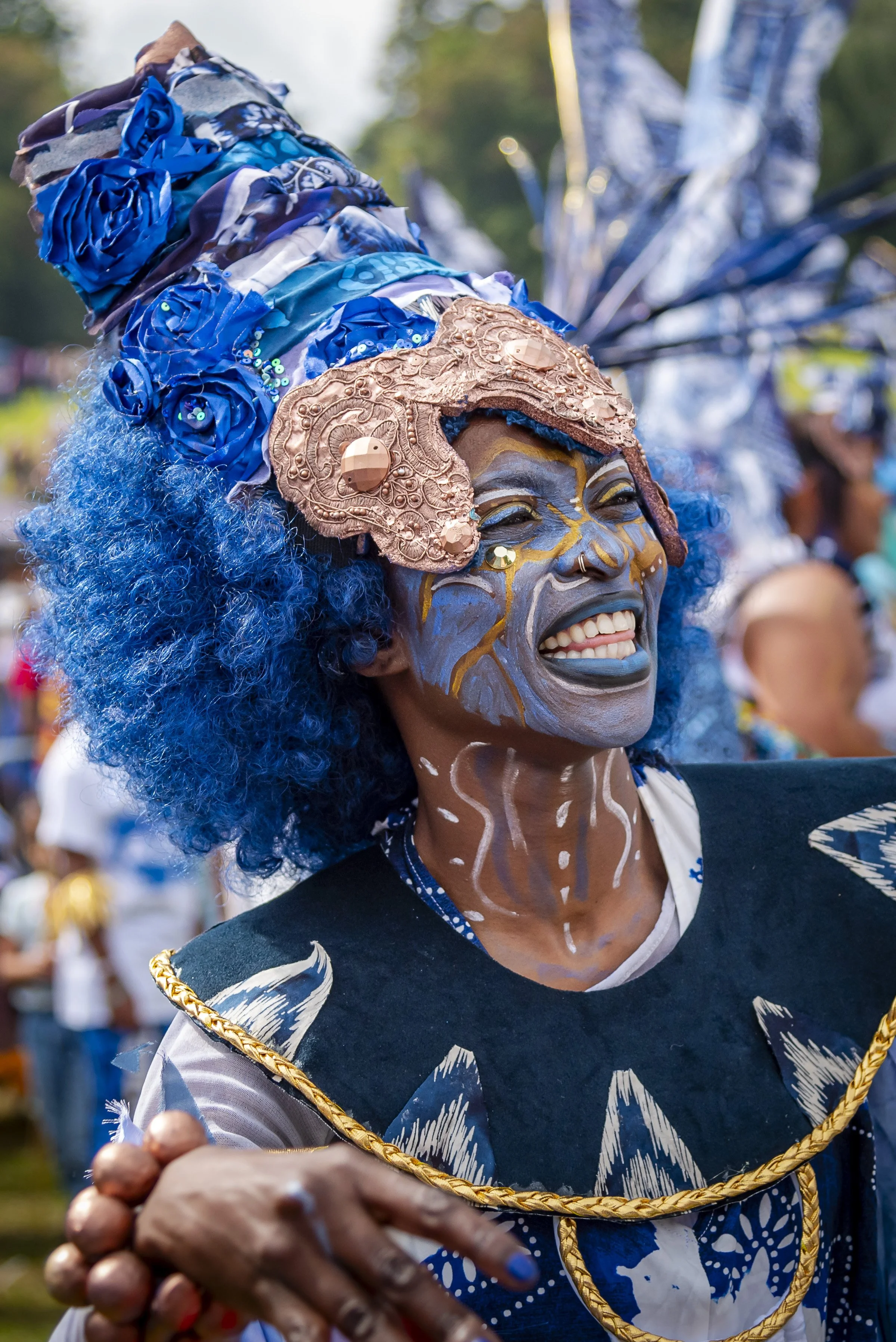 Person with blue curly hair, face painted with blue and yellow tribal patterns, wearing a headdress with blue fabric, sequins, and a large copper ornament, smiling at a celebration or parade.