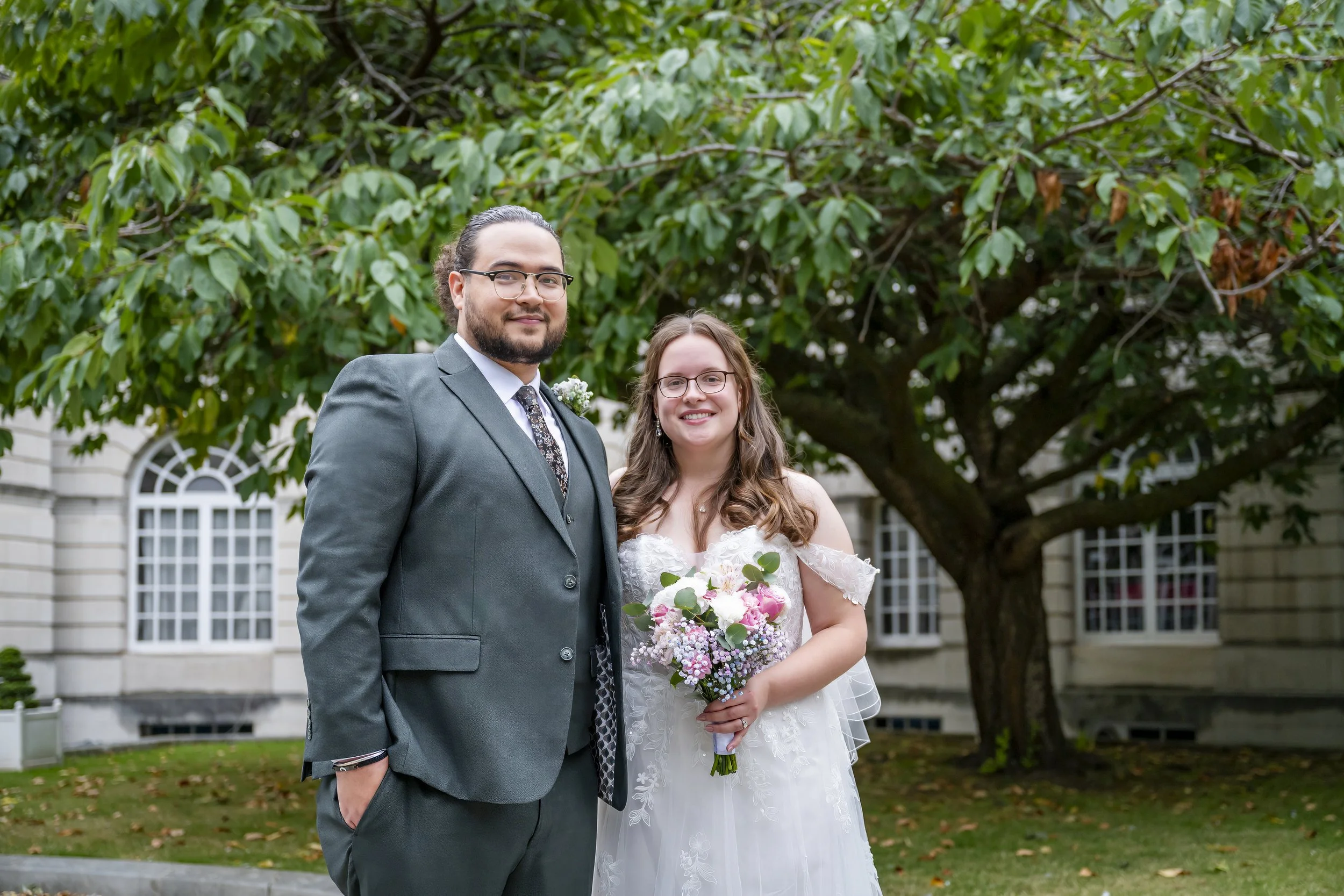 A newlywed couple standing outdoors in front of a large tree and historic building, with the groom in a gray suit and the bride in a white wedding dress holding a bouquet of flowers.