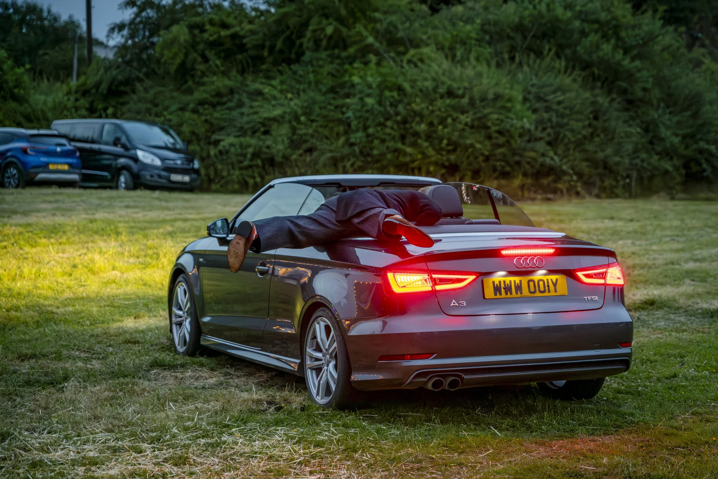 A man dressed in a suit lying on the back of a black Audi A3 convertible with a yellow license plate, in a grassy field with cars parked in the background.