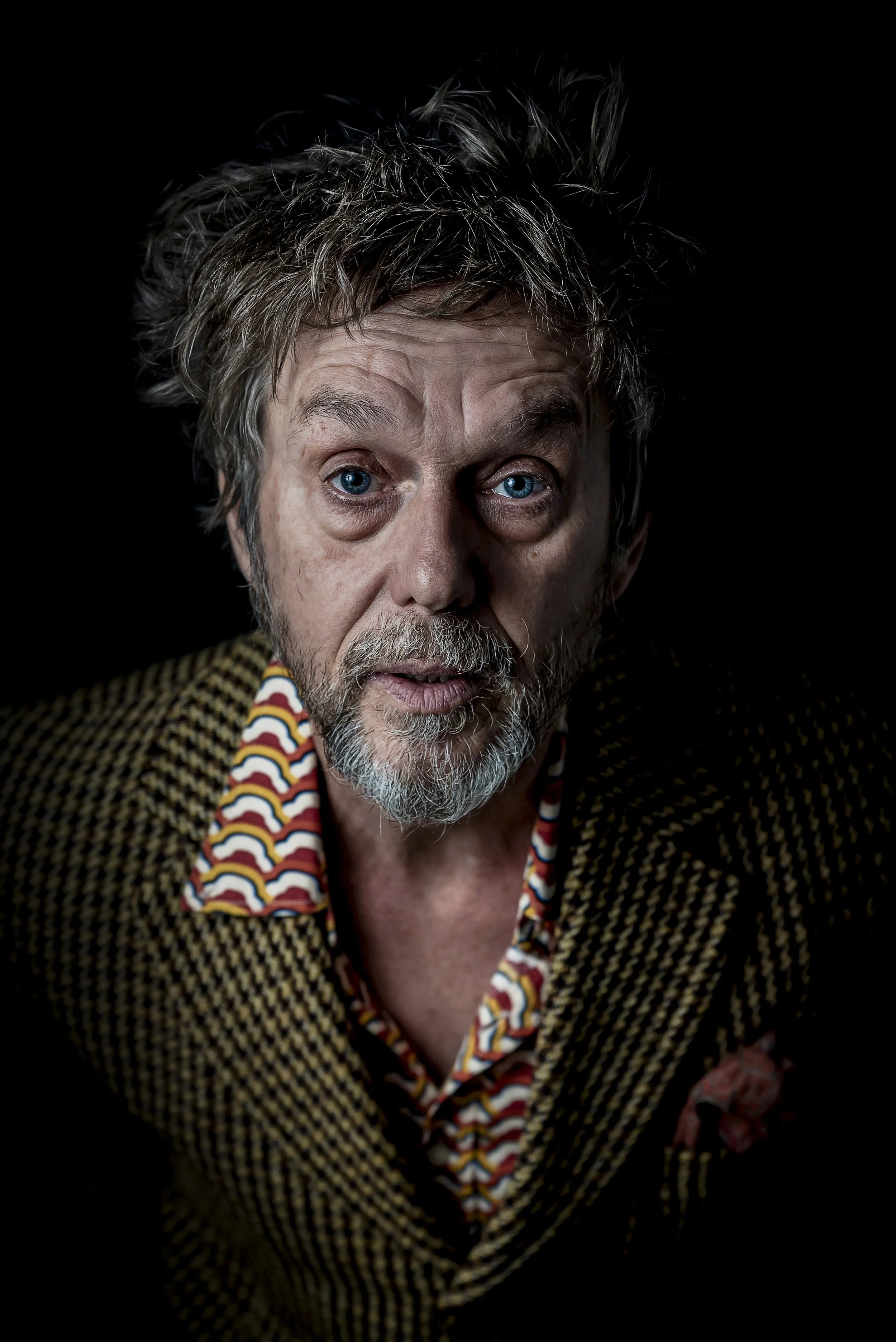 Portrait of an older man with disheveled gray hair and a beard, wearing a brown and yellow checked blazer over a patterned shirt, against a black background.