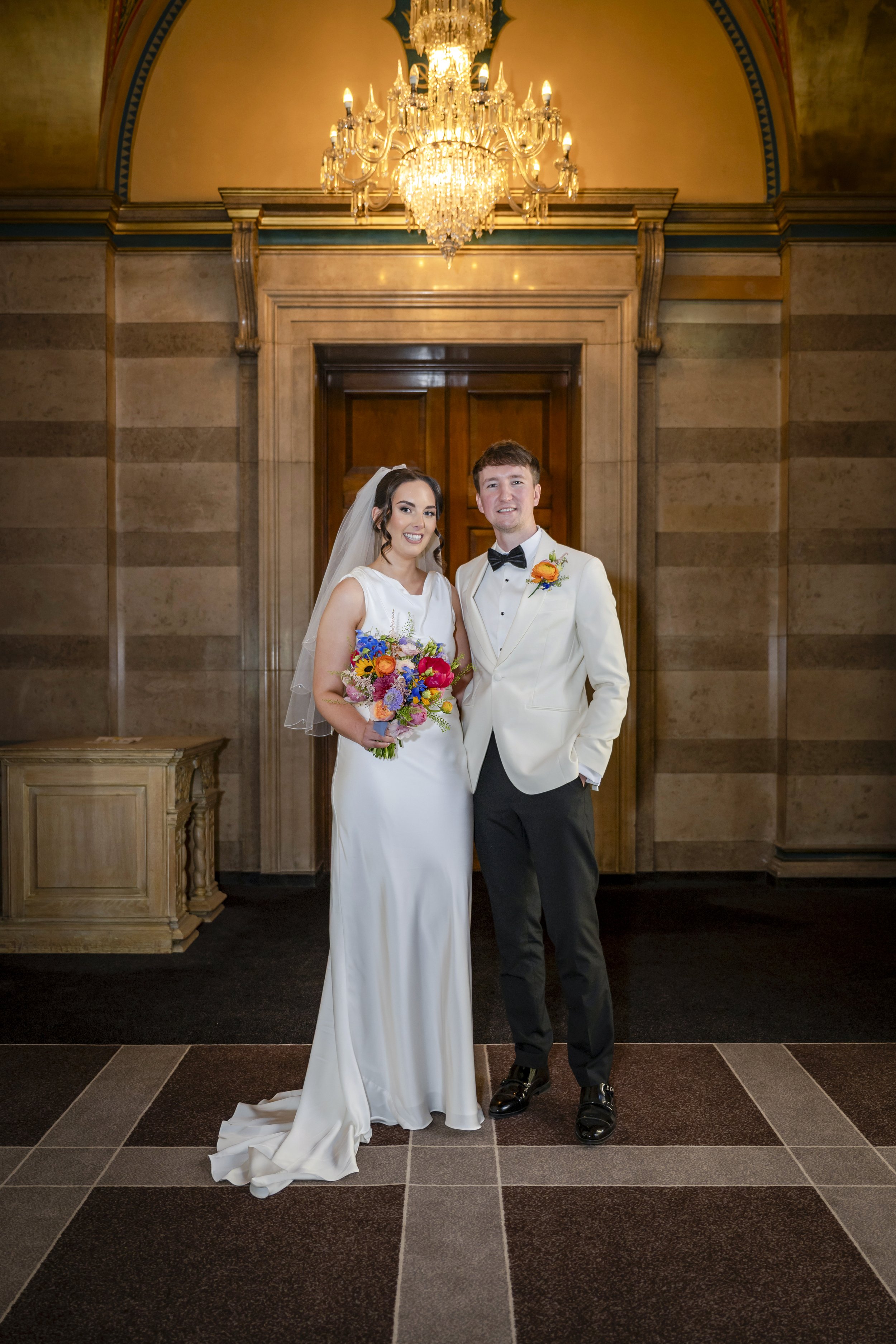 A bride and groom standing together in formal wedding attire in front of a wooden door with a grand chandelier hanging above them. The bride is holding a colorful bouquet of flowers and wearing a white wedding dress with a veil, while the groom is dr