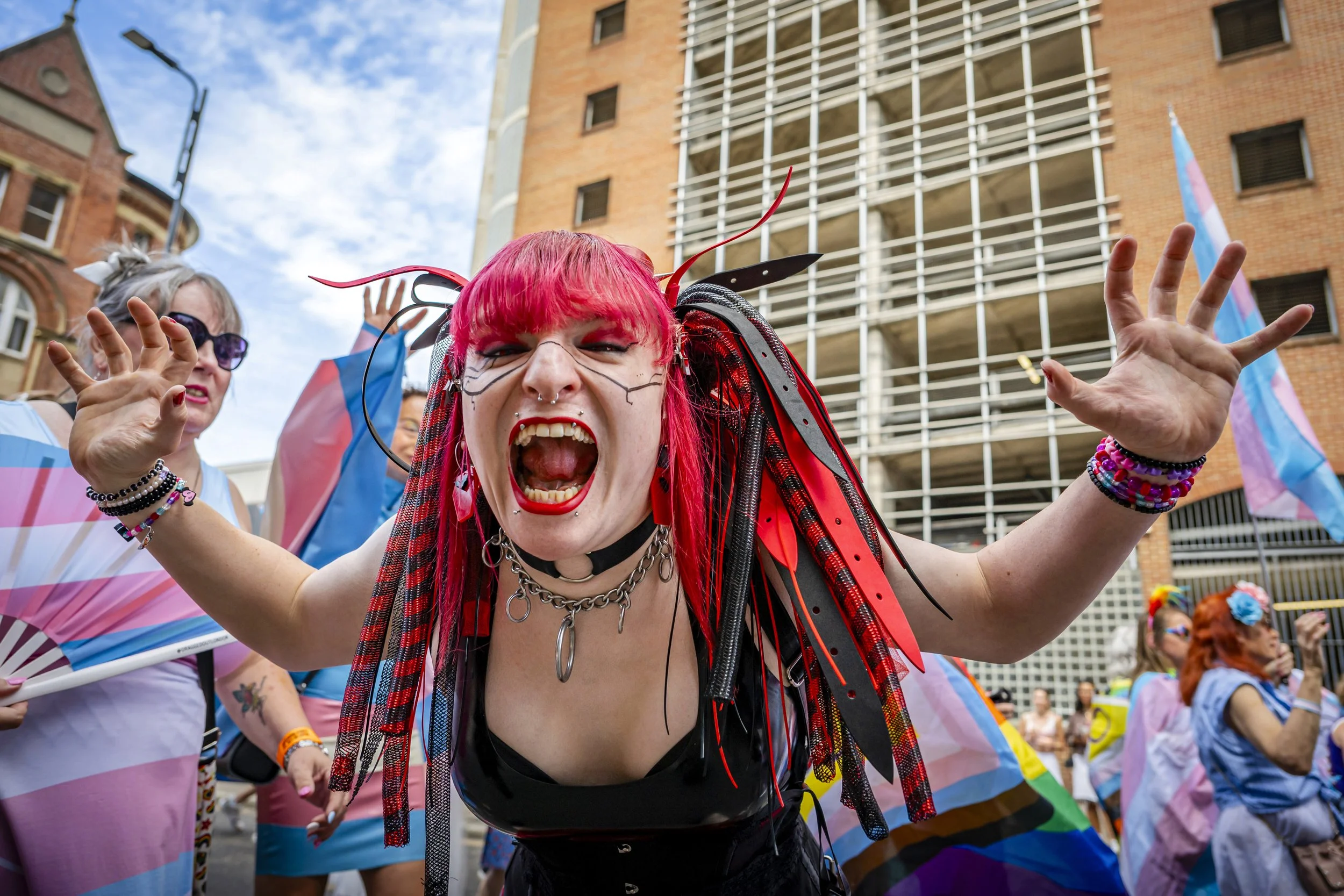 Person with bright pink hair, facial piercings, and face paint, shouting with arms raised at pride parade, with others and buildings in the background.