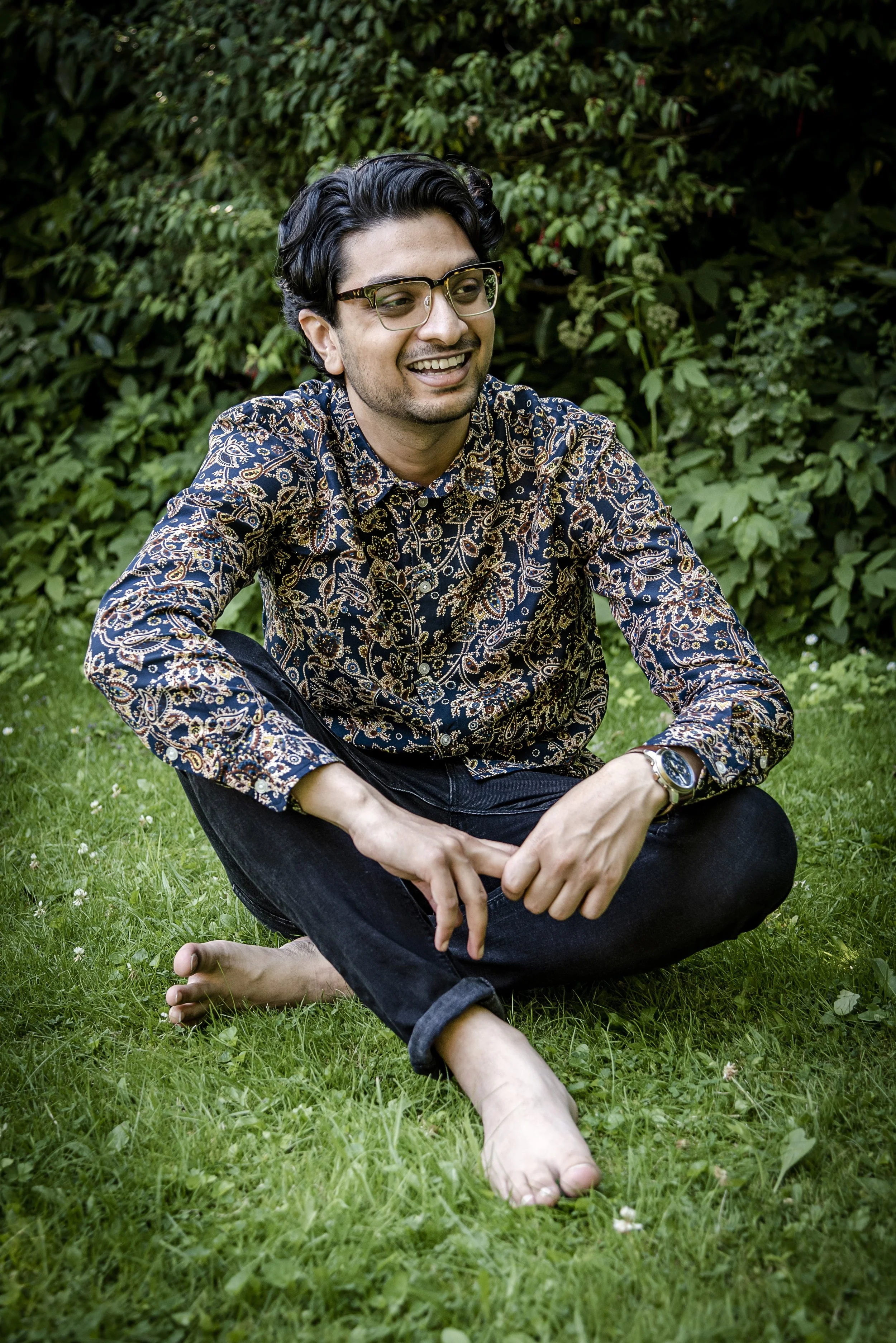 A man sitting on the grass outdoors, smiling, wearing glasses, a patterned shirt, black pants, and a watch, with greenery in the background.