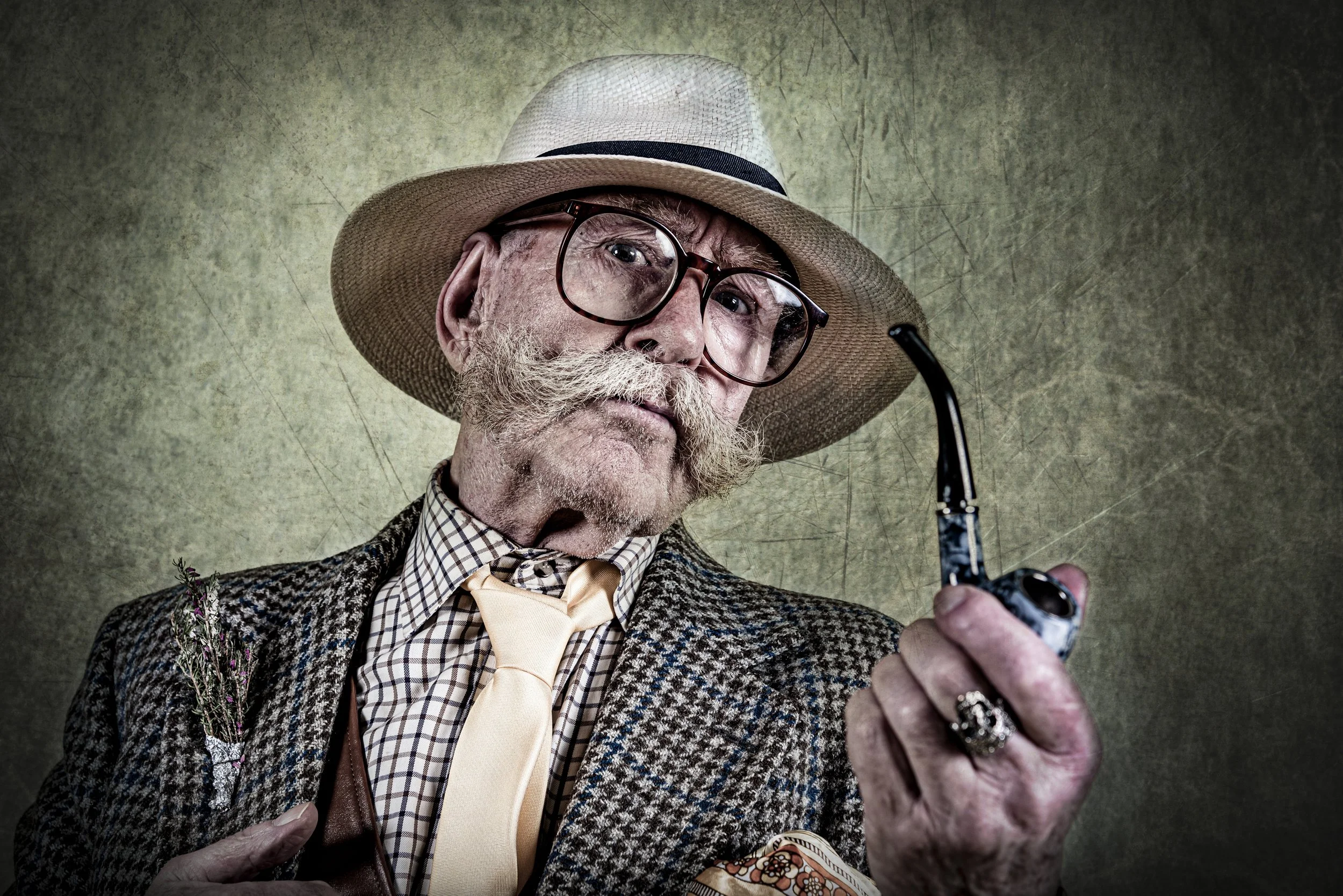 An elderly man with a white mustache and beard wearing a plaid jacket, a beige tie, and a fedora hat holding a smoking pipe. He has glasses and a flower pinned to his jacket, posing against a textured green background.
