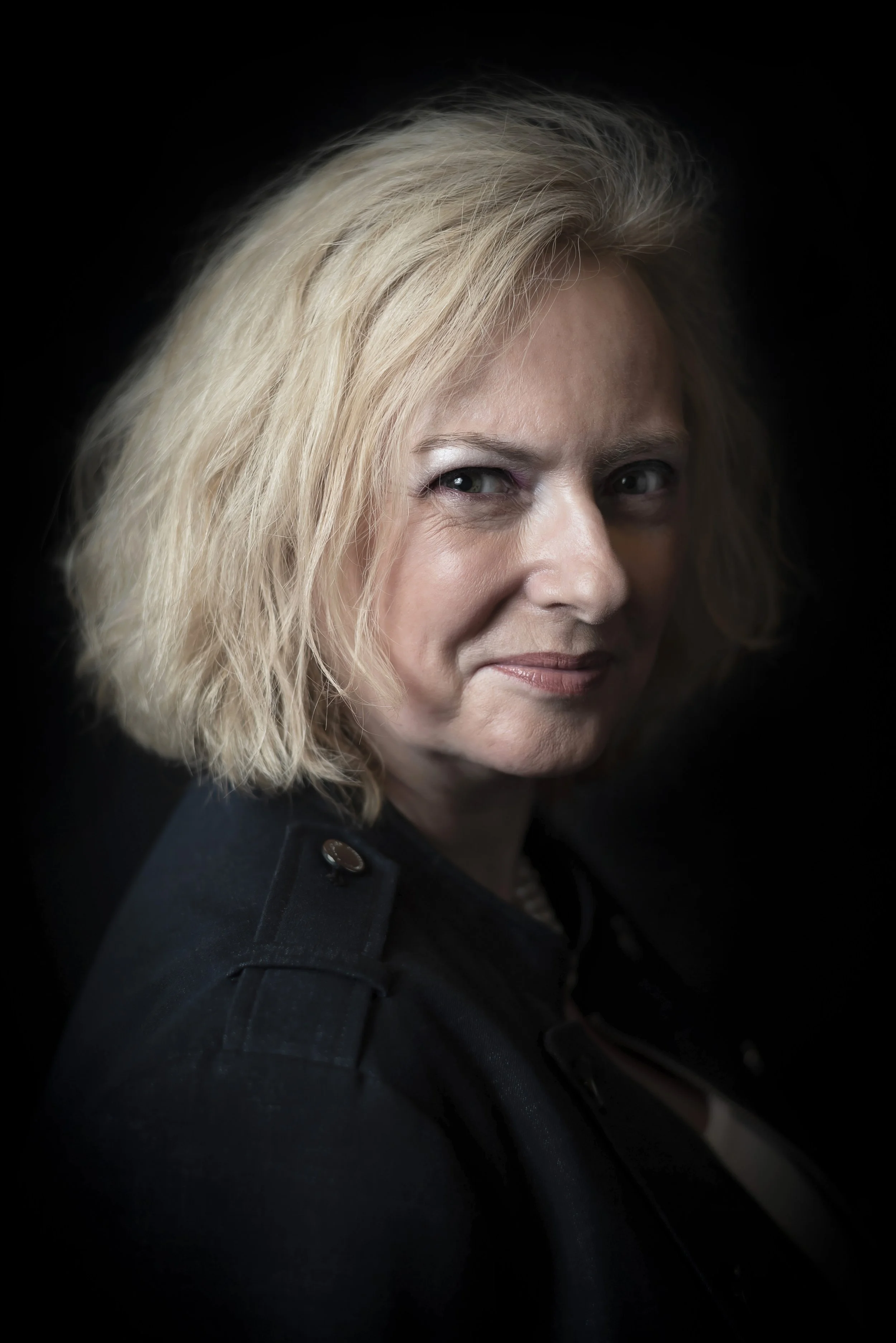 Portrait of a middle-aged woman with blonde hair, wearing a dark jacket, against a black background.