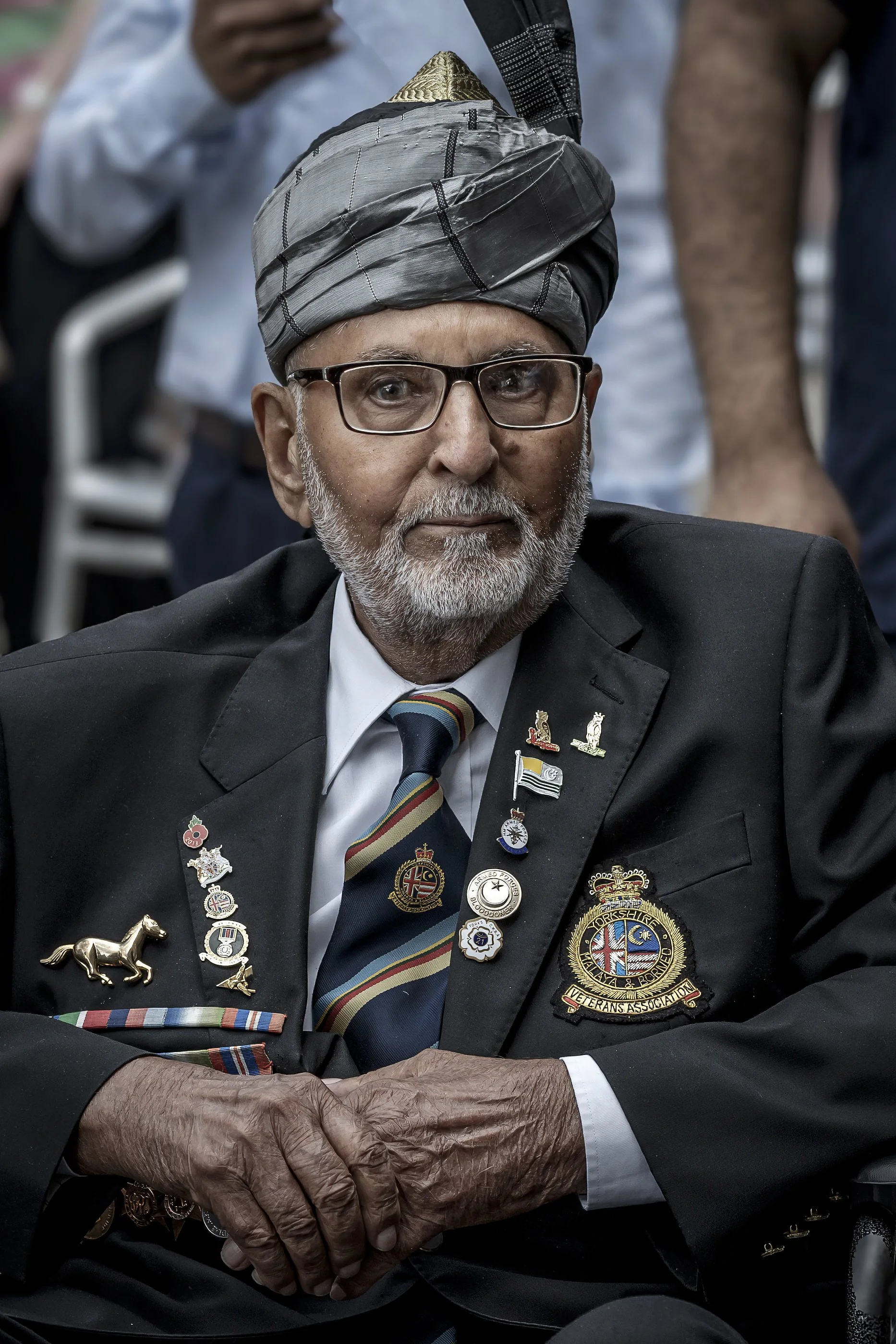 An elderly man wearing a traditional turban, glasses, a black suit jacket with numerous pins and badges, a striped tie, and a dress shirt, sitting at a formal event.