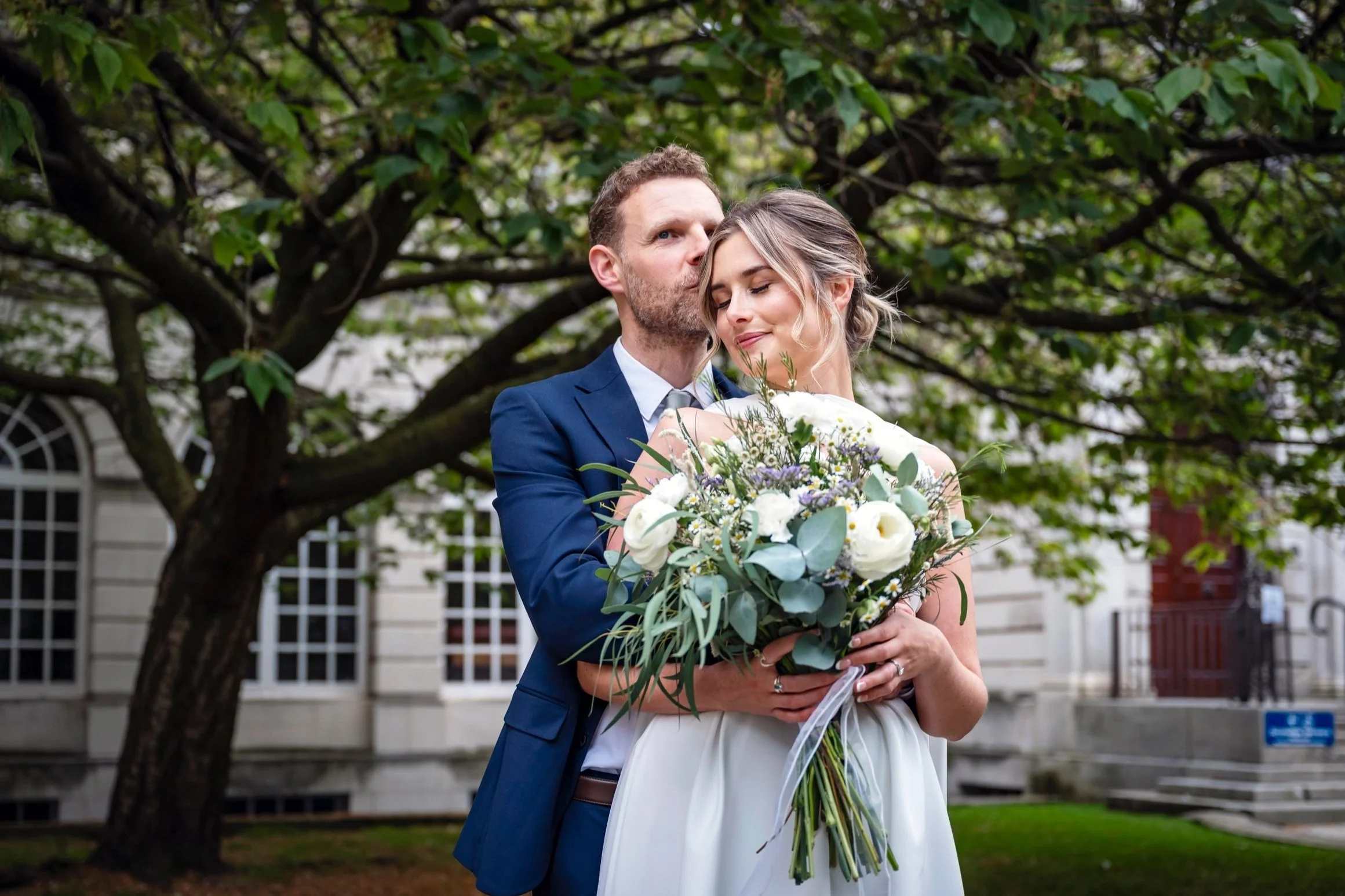 A bride and groom embrace outdoors under a large green tree, the groom kissing the bride's forehead while she holds a bouquet of white and purple flowers, with visible historic building in the background.