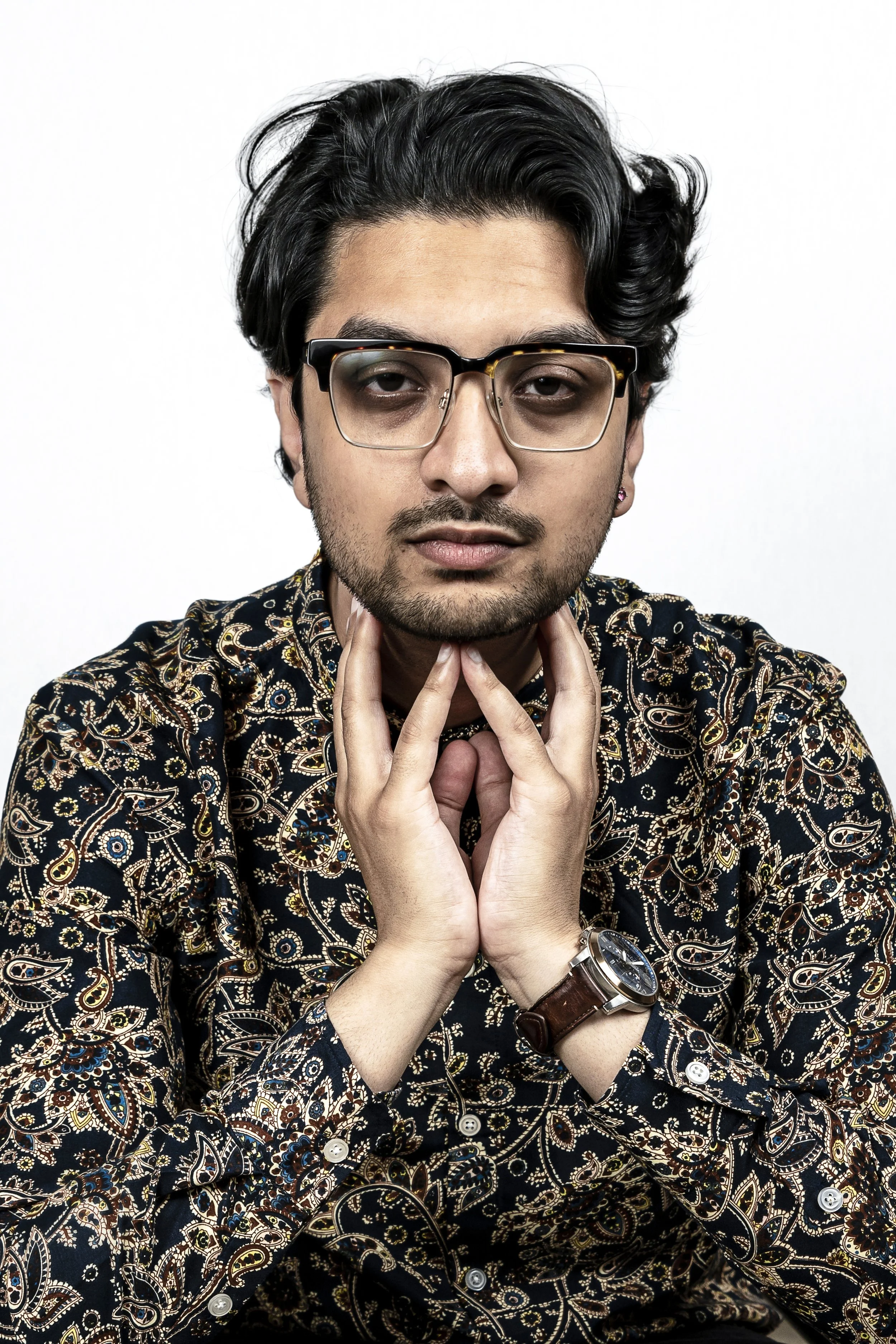 A man with black hair, wearing glasses, a patterned shirt, and a wristwatch, looking directly at the camera with his hands gently touching his neck.