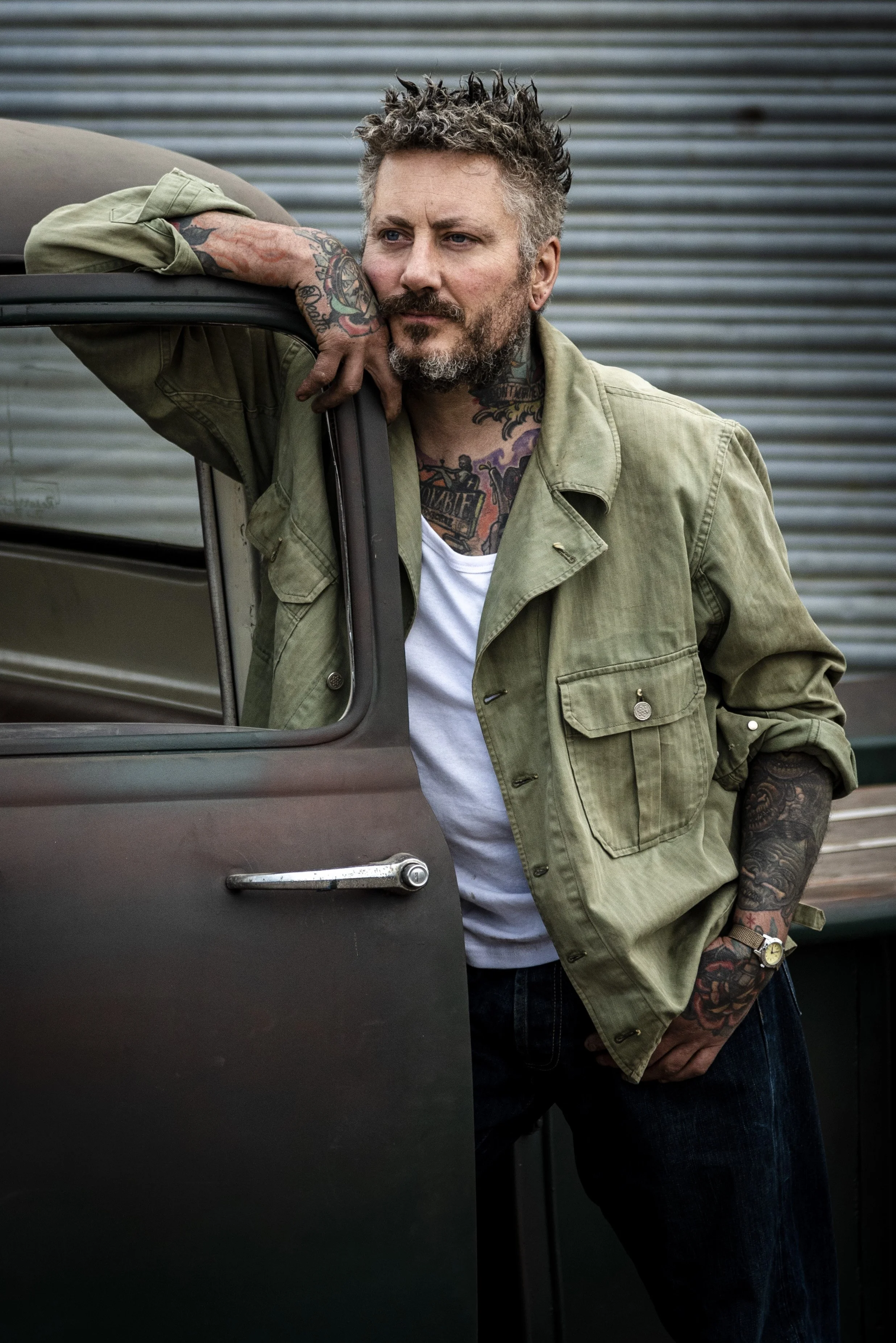 A man with tattoos, styled hair, and a beard leaning on a truck with a relaxed pose, outdoors against a metal building background.