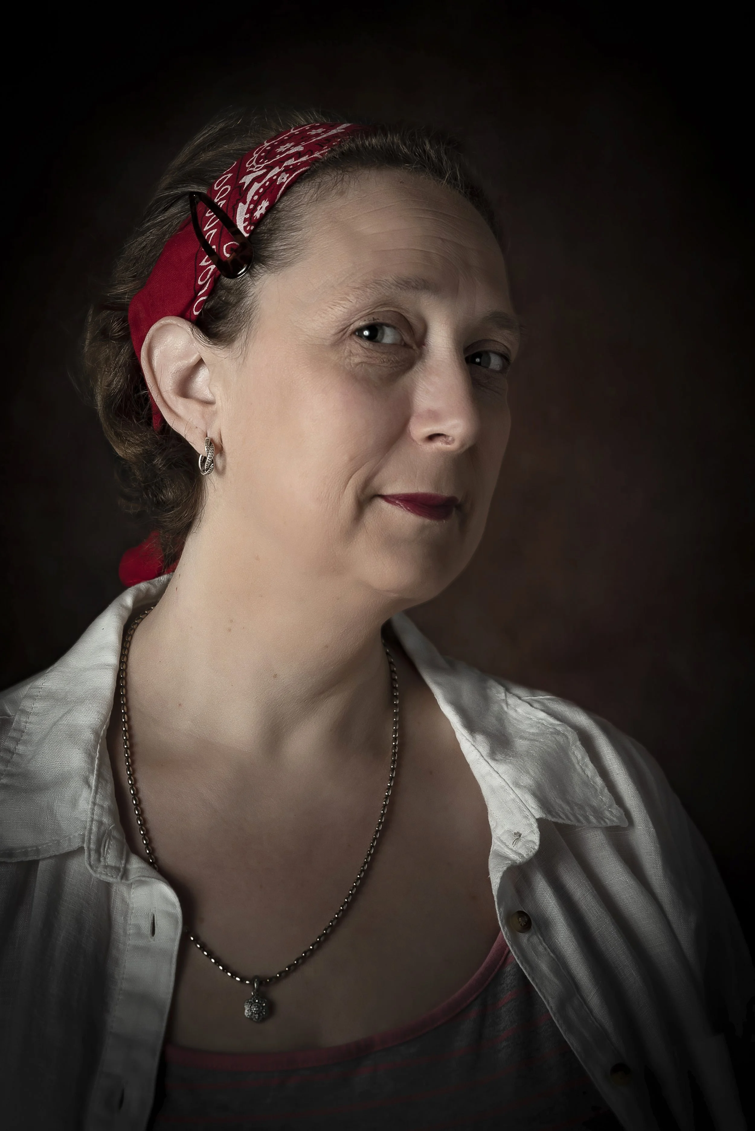 Portrait of a woman with a red bandana in her hair, wearing earrings and a necklace, and dressed in a white shirt and striped top, against a dark background.