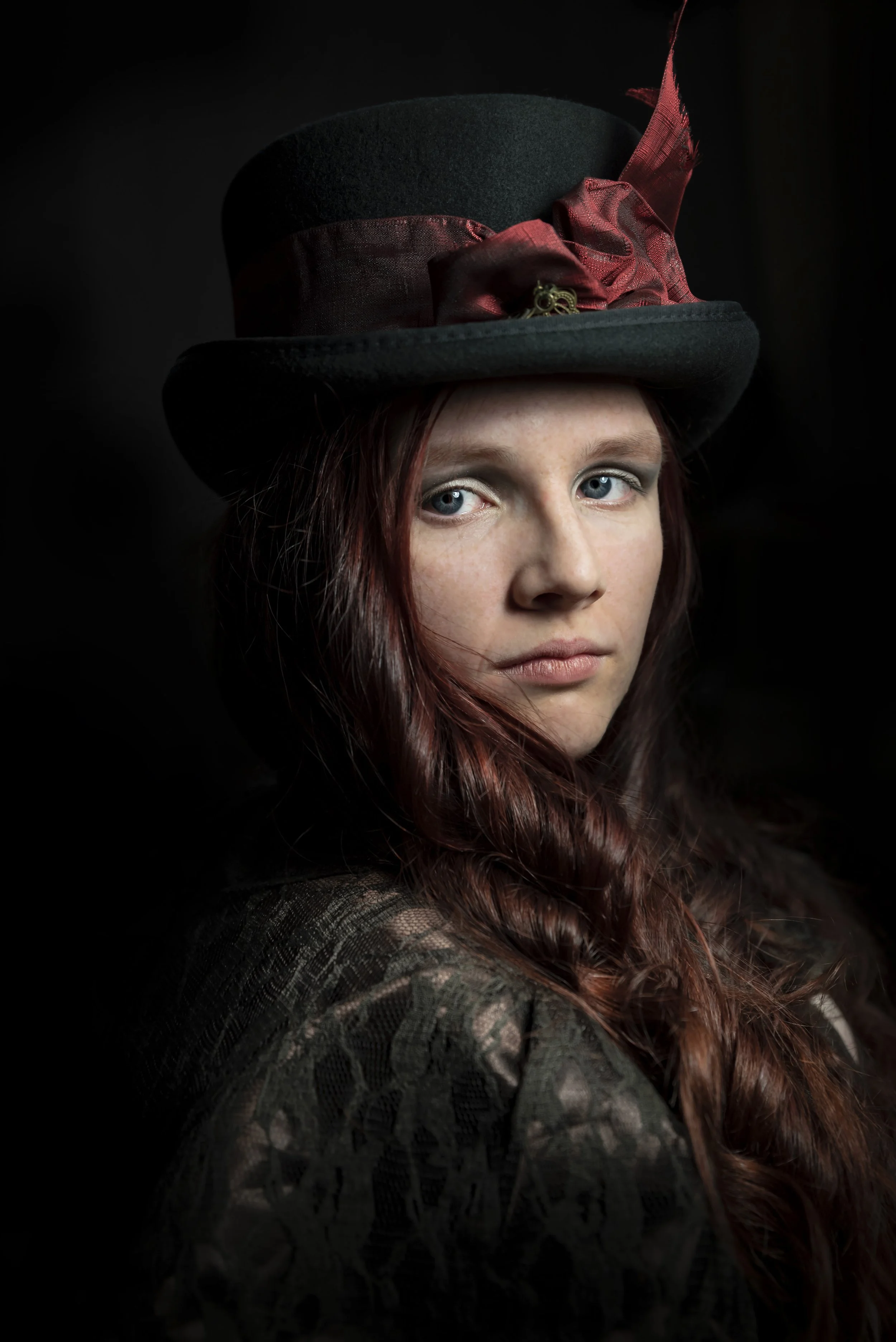 A young woman with red hair and blue eyes wearing a vintage black top hat with a red ribbon and feather, and a patterned black lace dress, against a dark background.