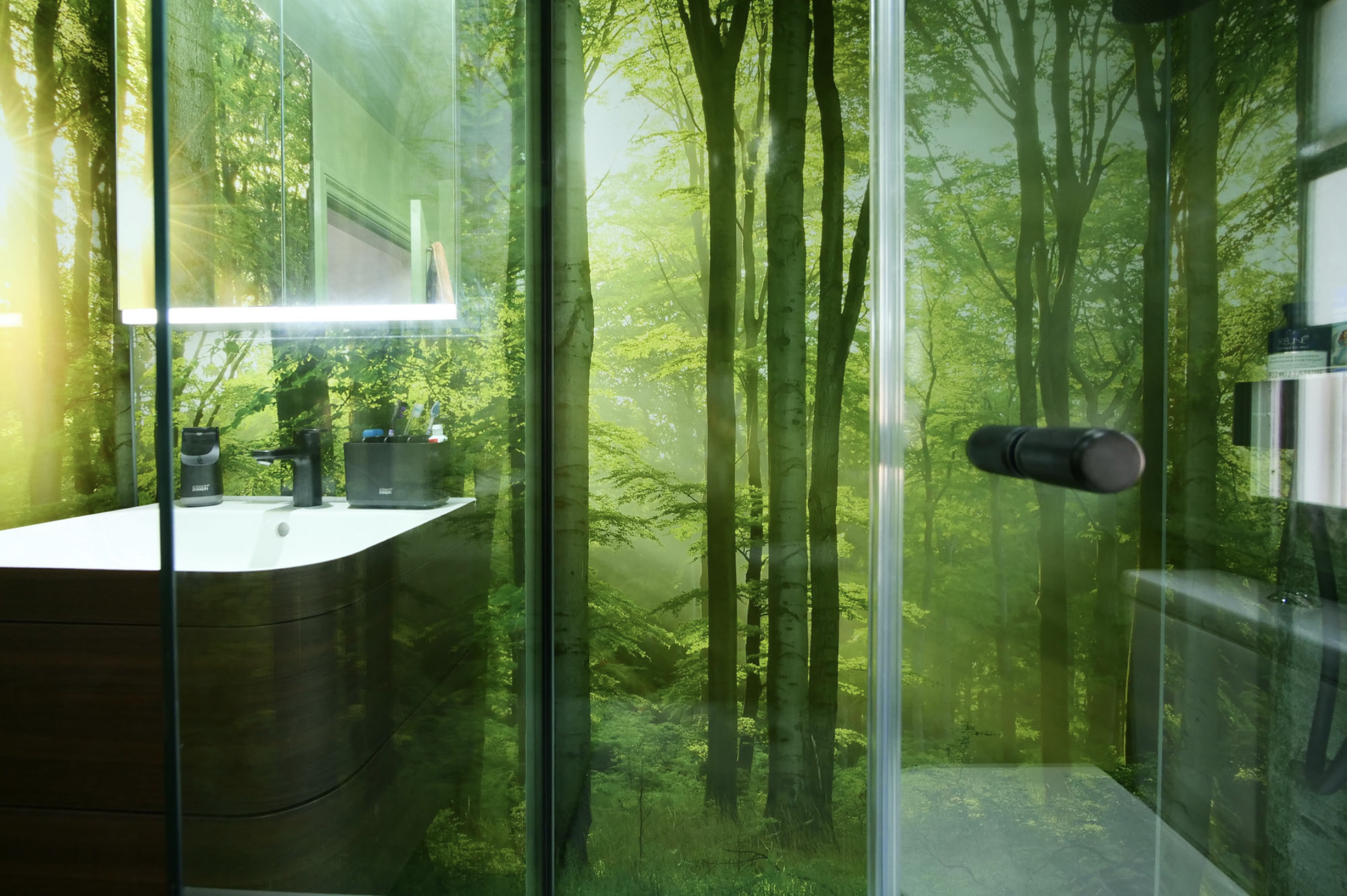 Photo of shower area of a modern bathroom with wrap-around wall panels with lush green forest imagery. sliding glass shower screen in foreground.