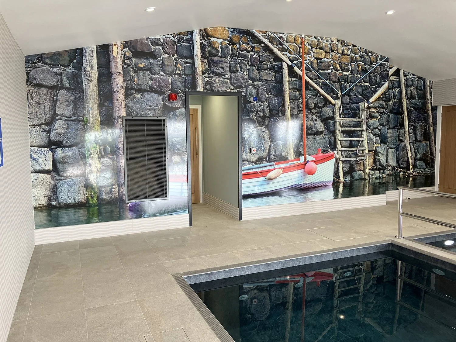 Indoor swimming pool with large feature wall image showing a stone harbour wall with red and white painted rowing boat.