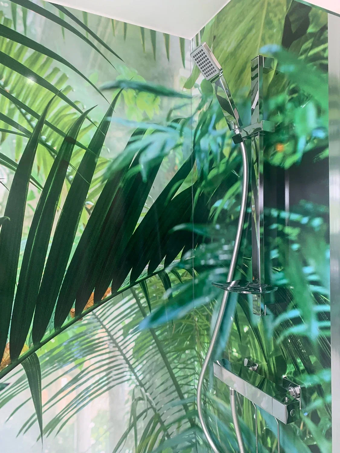 Customer installation photo of Jungle Mist design shower wall panels wrapping around a corner shower, with lush green tropical plants reflected in chrome shower fittings.