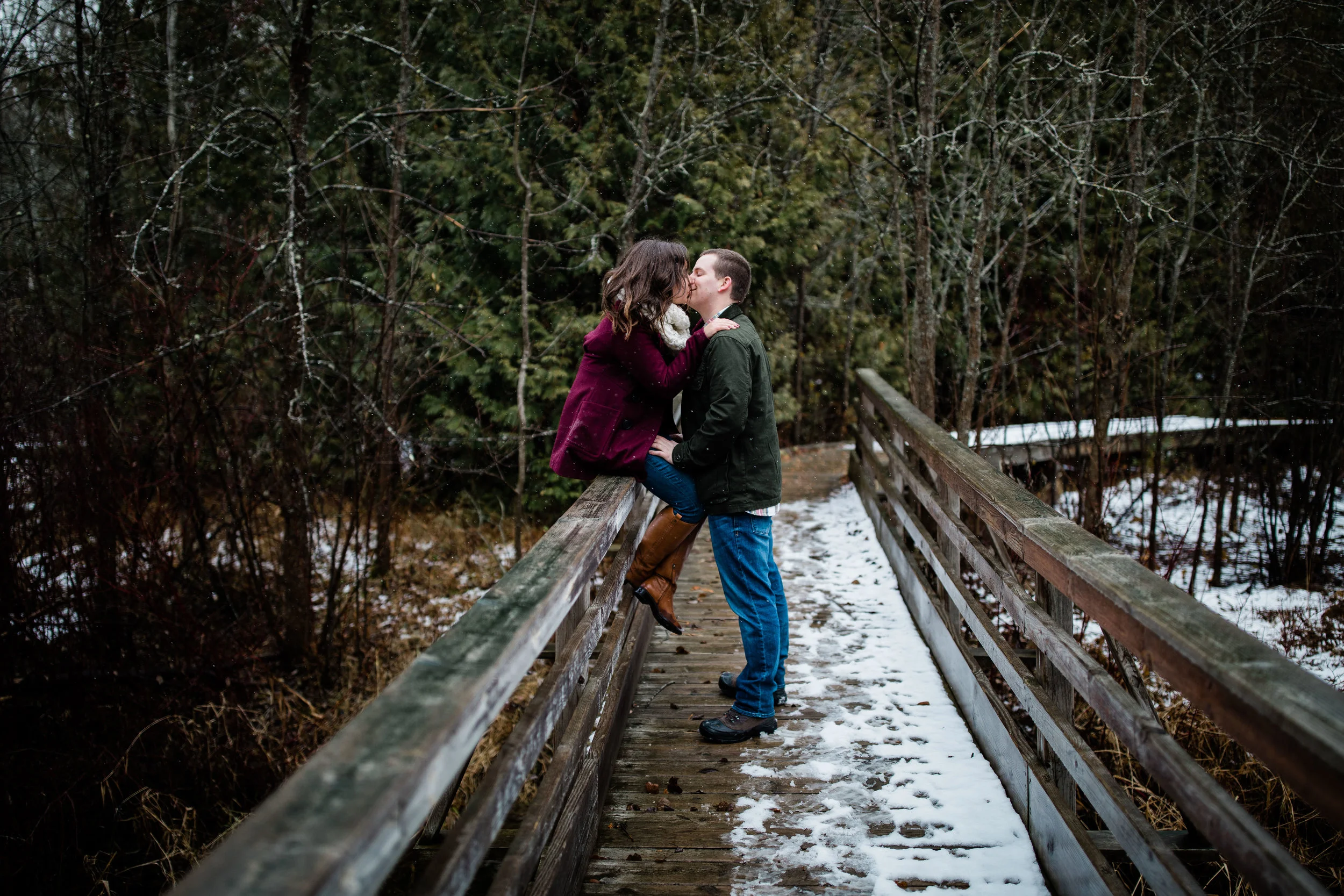 Libby sits on a wooden footbridge railing and kisses Alex. Snow is on the ground.