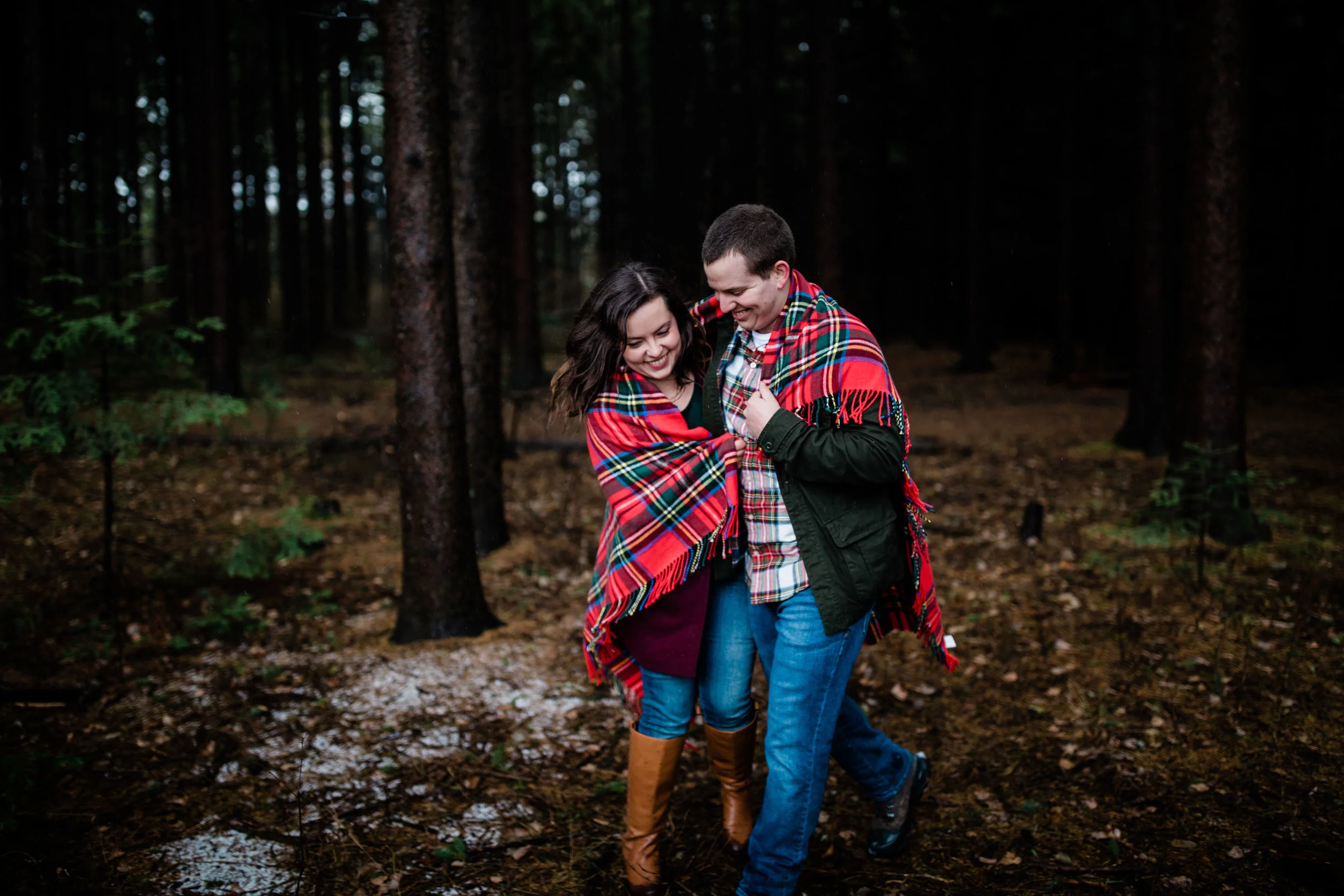 Libby and Alex are both wrapped in a red blanket walking through the woods.