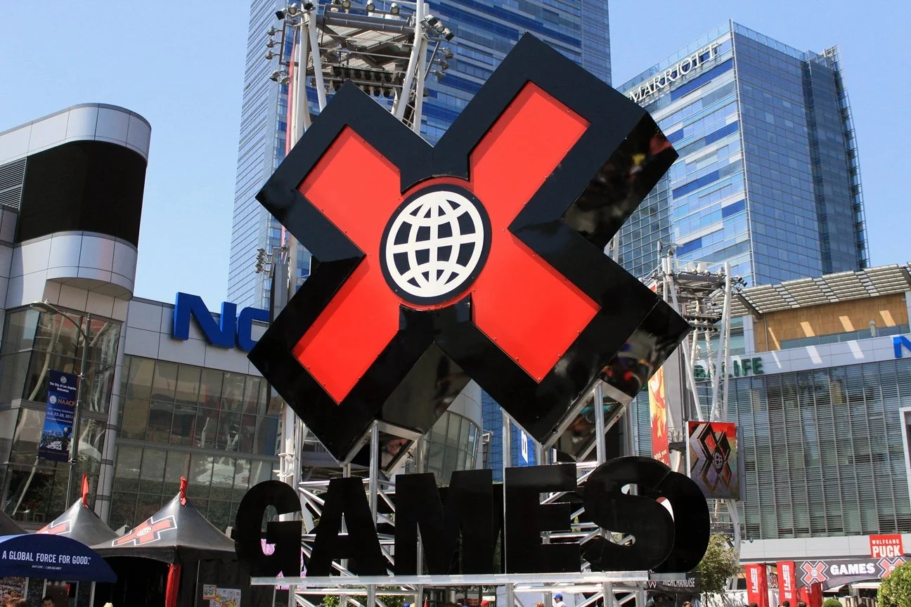 Large X Games sign with a stylized globe, set in a cityscape with modern buildings and structures, during daytime.