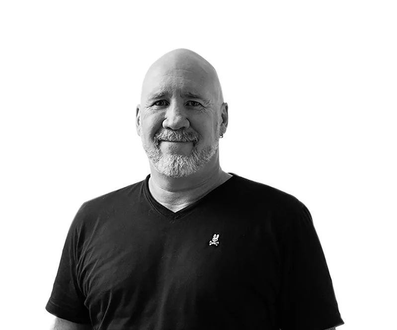 Black and white portrait of a bald man with a beard, wearing a black V-neck T-shirt, smiling, against a plain white background.
