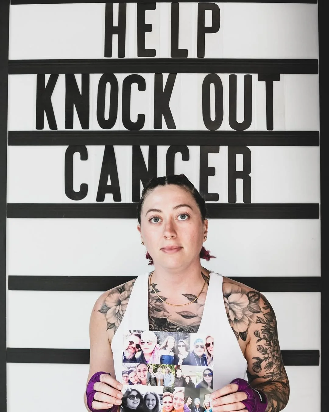 A woman with tattoos holding a collage of photos, standing in front of a sign that reads 'Help Knock Out Cancer'.