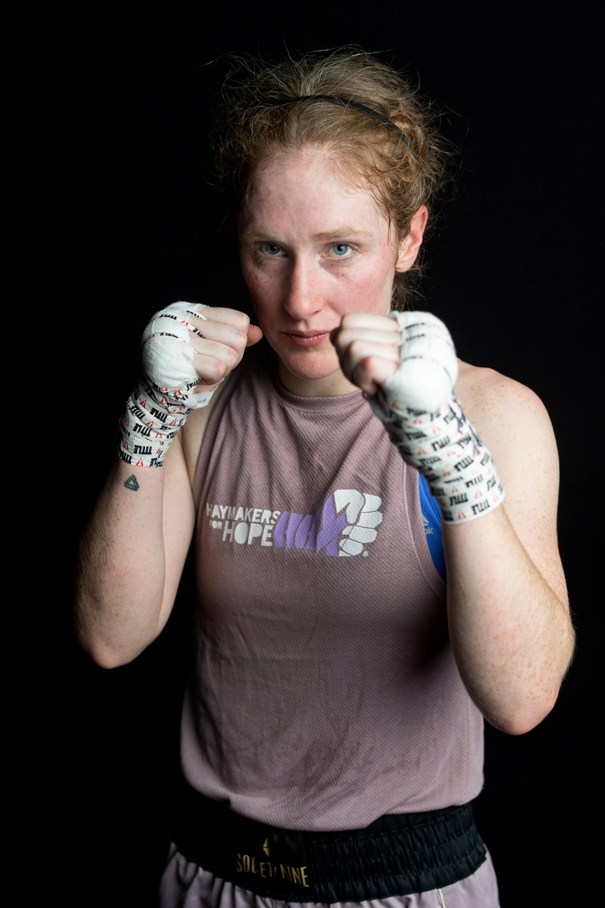 Female boxer in a fighting stance, wearing hand wraps and athletic gear, against a black background.