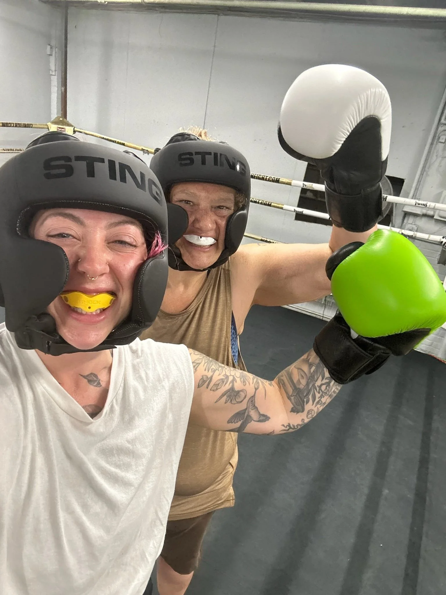 Two women wearing boxing helmets and gloves are happy and posing for a selfie in a gym. One woman is holding up her arm with a boxing glove, and they are smiling.