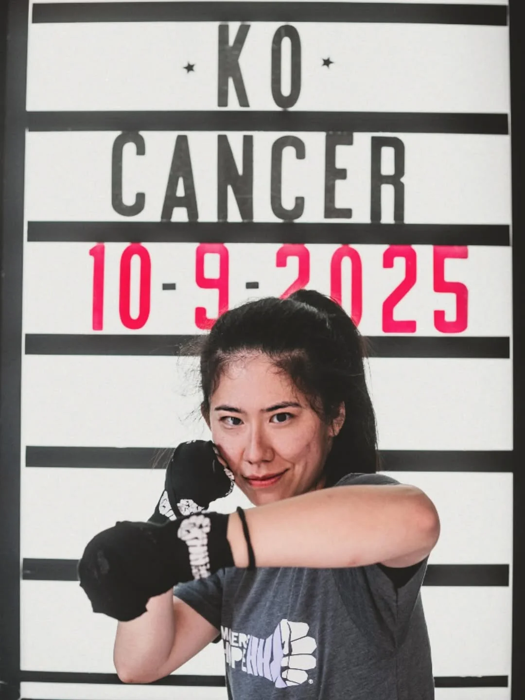 A woman in a workout shirt and gloves poses with her arm raised in front of a height measurement board with a mugshot style sign. The sign displays the date 10-9-2025 and the words 'K O CANCER' with black, grey, and pink text.