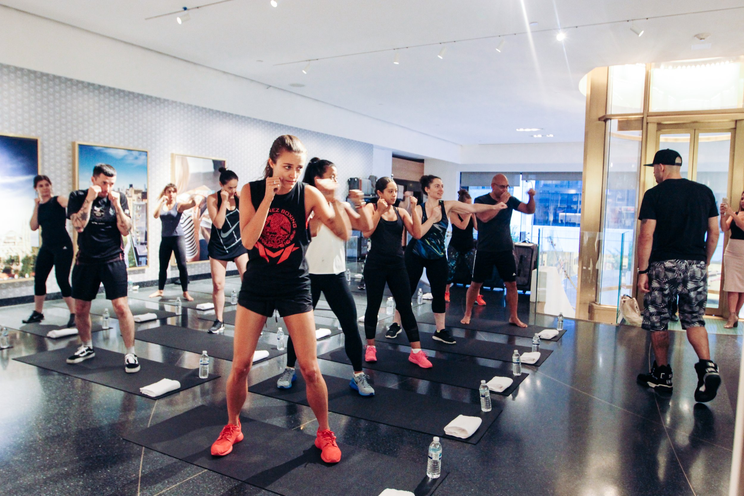 Group of people participating in a fitness class with instructor leading, all standing on black mats with water bottles and towels, inside a bright room with large windows and artwork on the walls.