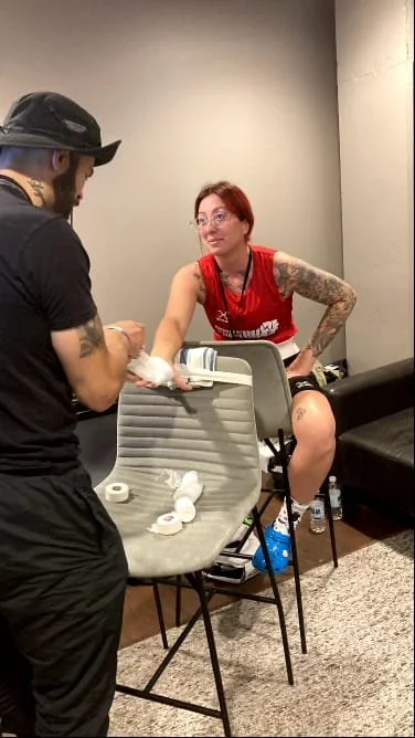 A woman with tattoos in a red sports jersey is sitting on a black chair, receiving a tattoo on her left leg from a tattoo artist in a black cap and shirt in a room with gray walls.