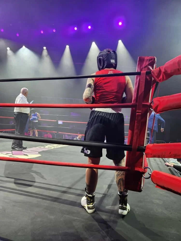 A boxer standing in a boxing ring, viewed from behind, wearing a red sleeveless top, black shorts, and headgear, with tattooed legs, inside an arena with purple and white stage lights.