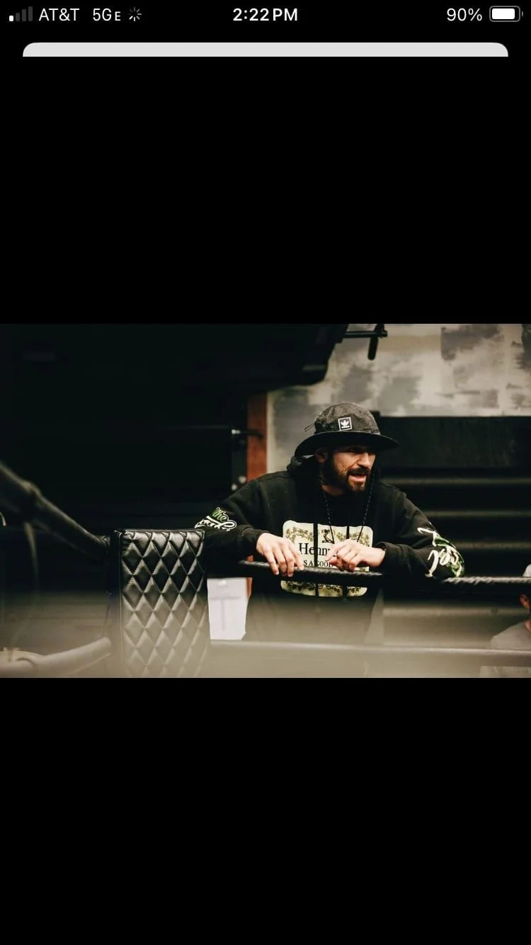 A man with a beard wearing a black hoodie and a black bucket hat with a logo, standing inside a boxing gym, leaning on the ropes of the boxing ring.