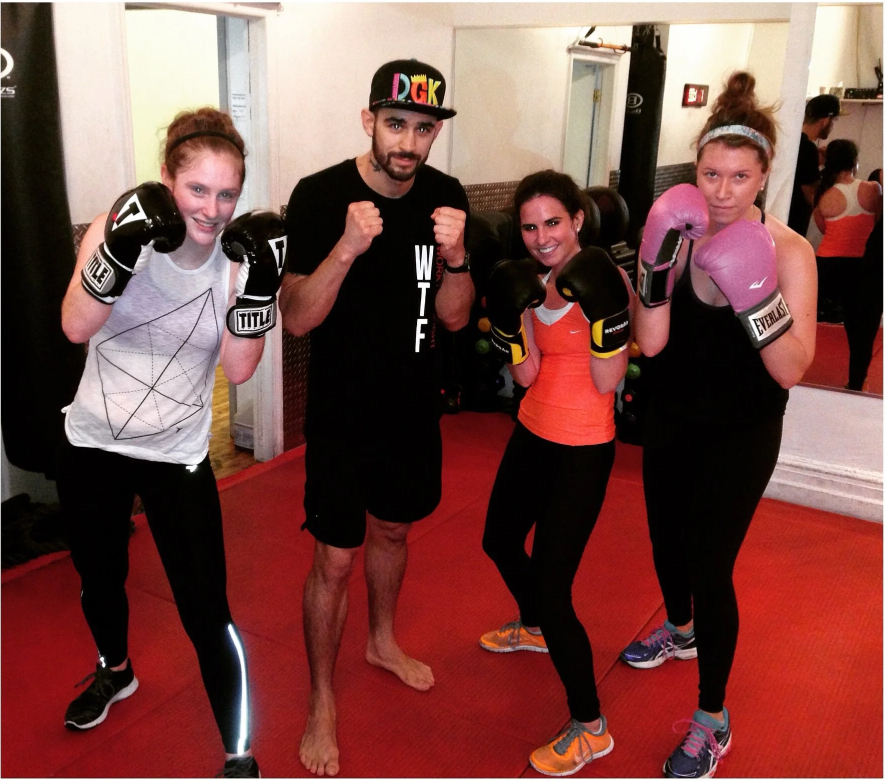 Four women and one man in boxing gear and athletic clothing posing together in a gym, all with their fists raised. The women are wearing boxing gloves, and the man is wearing a cap and shorts. They are standing on a red mat in front of a mirror.