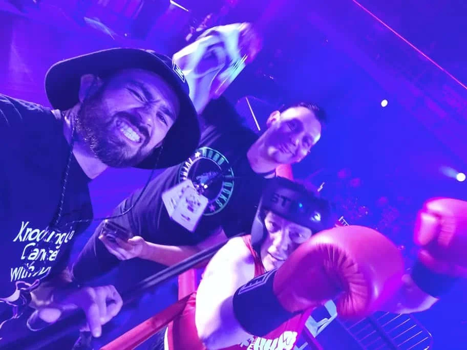 Three people inside a boxing ring, two men and one woman, posing for a photo; the woman is wearing red boxing gloves and headgear, and the men are smiling, with one giving a thumbs-up. The scene has purple and pink lighting, suggesting a lively envir