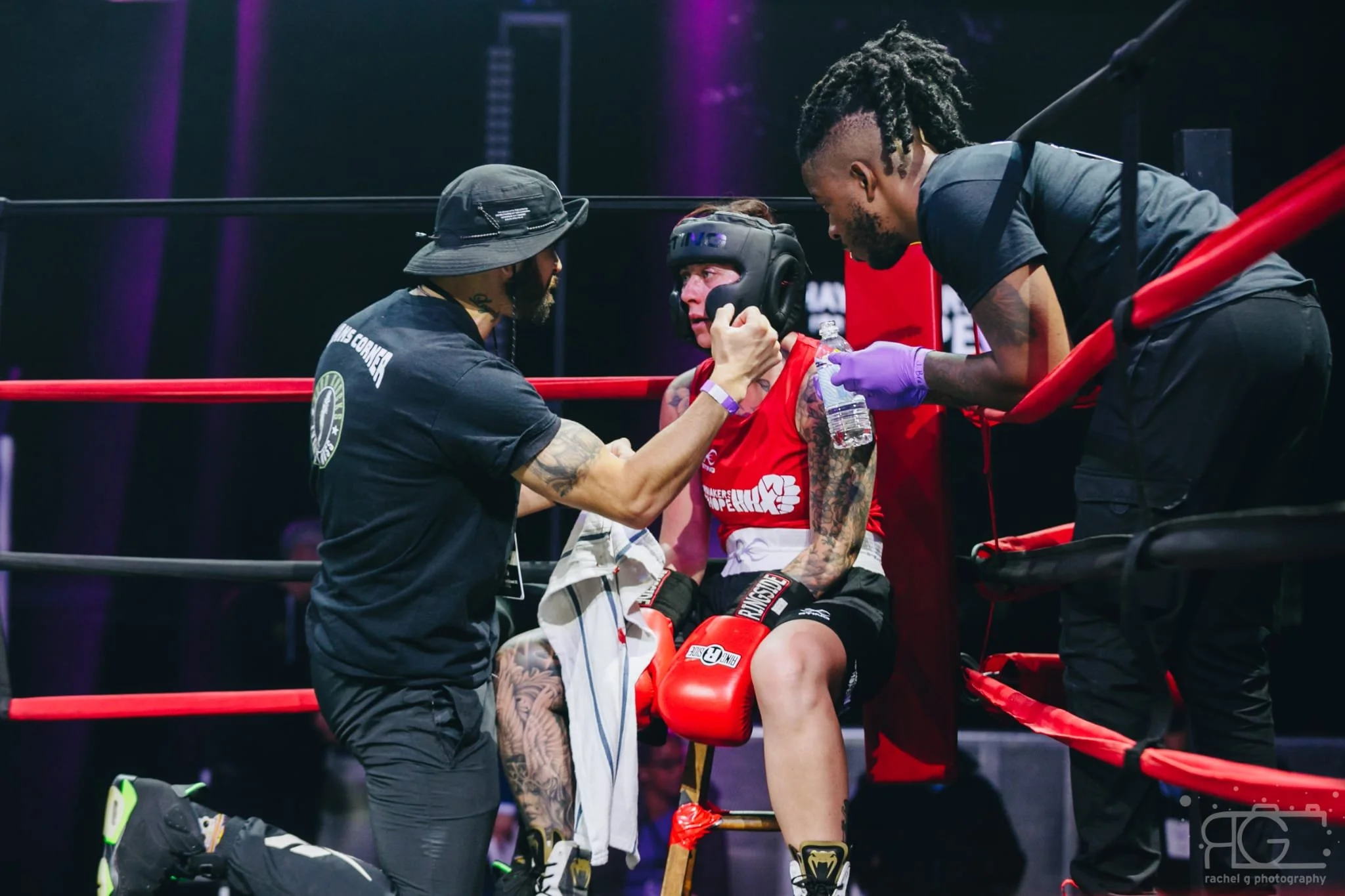 A female boxer sitting in her corner of the boxing ring, receiving water and encouragement from two team members, one holding her chin and the other offering water while she wears boxing gloves, shorts, and headgear.