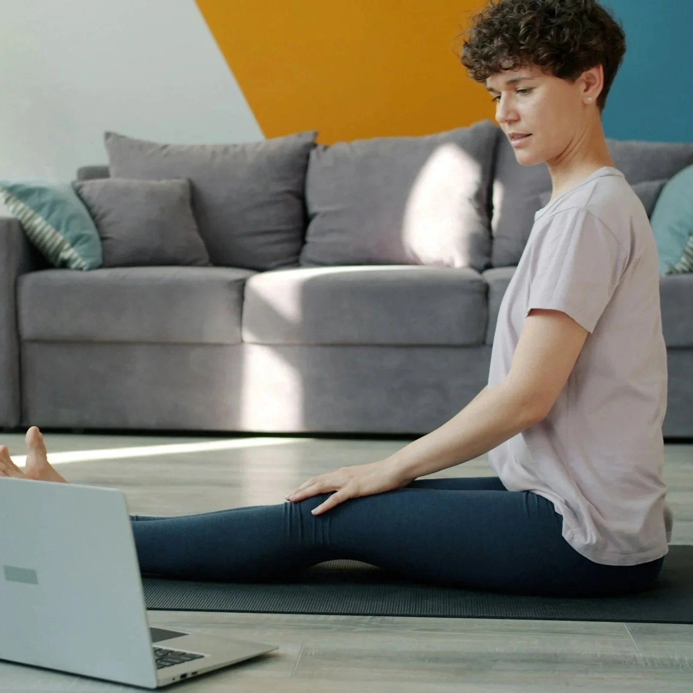 A woman sitting on a yoga mat in a living room, stretching her leg while looking at a laptop.