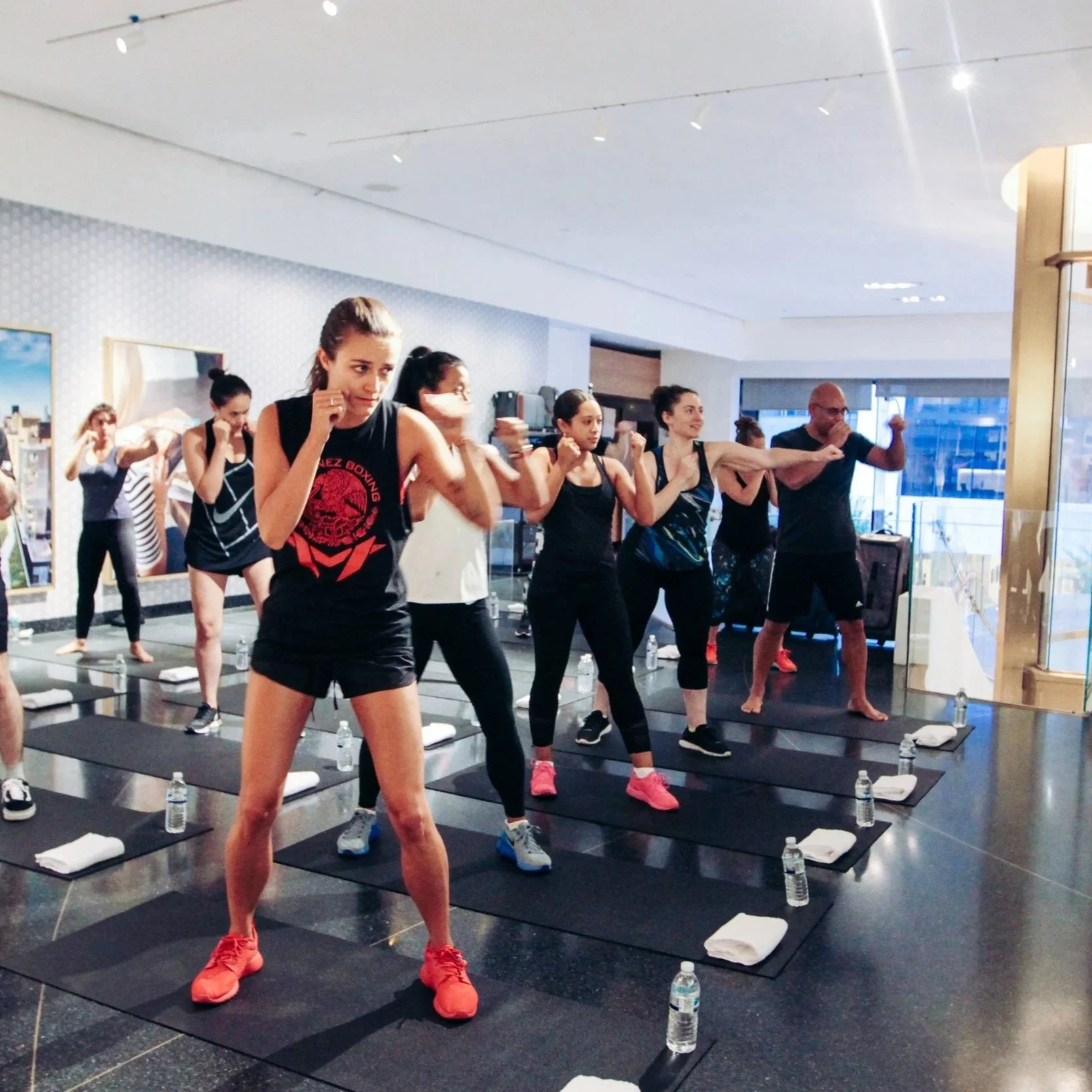 A group of women and men participating in a fitness or boxing class in a gym or studio, wearing workout attire, standing on black mats, with water bottles and towels nearby, in a well-lit modern space.