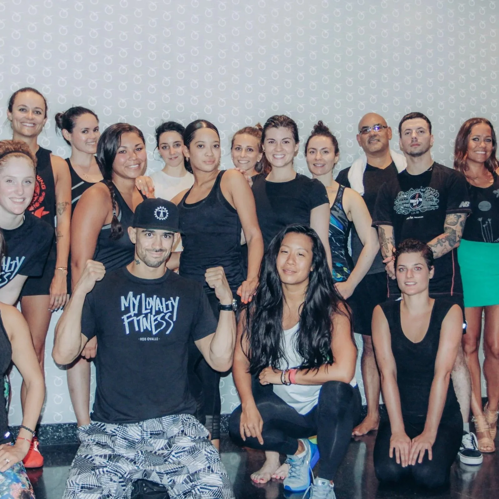 Group of people, mostly women, in athletic clothing posing together indoors, some smiling, one man in front with raised fists wearing a black cap and t-shirt that reads 'My Loyalty Fitness'.