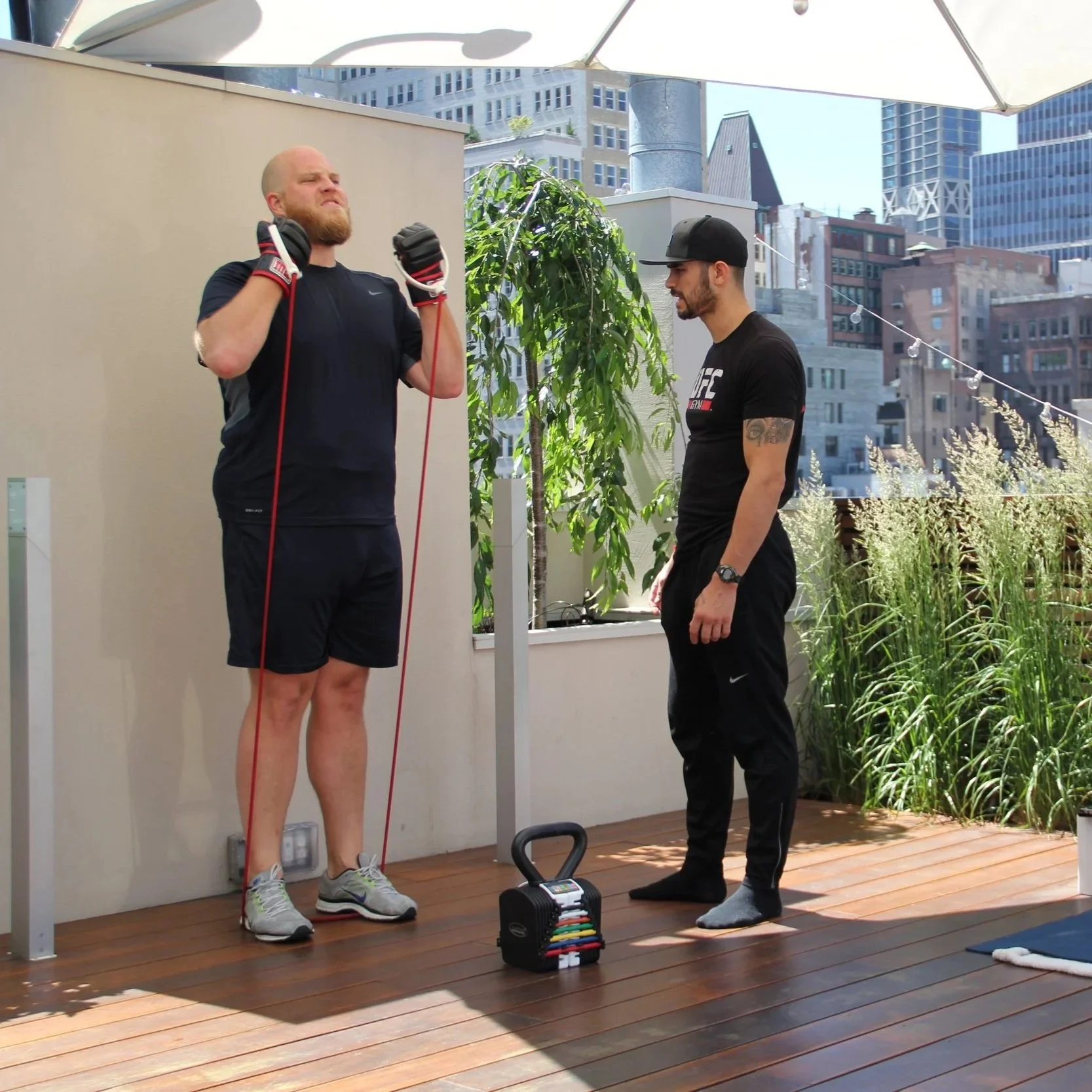 Two men on a rooftop patio, one stretching with resistance bands, the other standing nearby in casual athletic wear, with city buildings in the background.