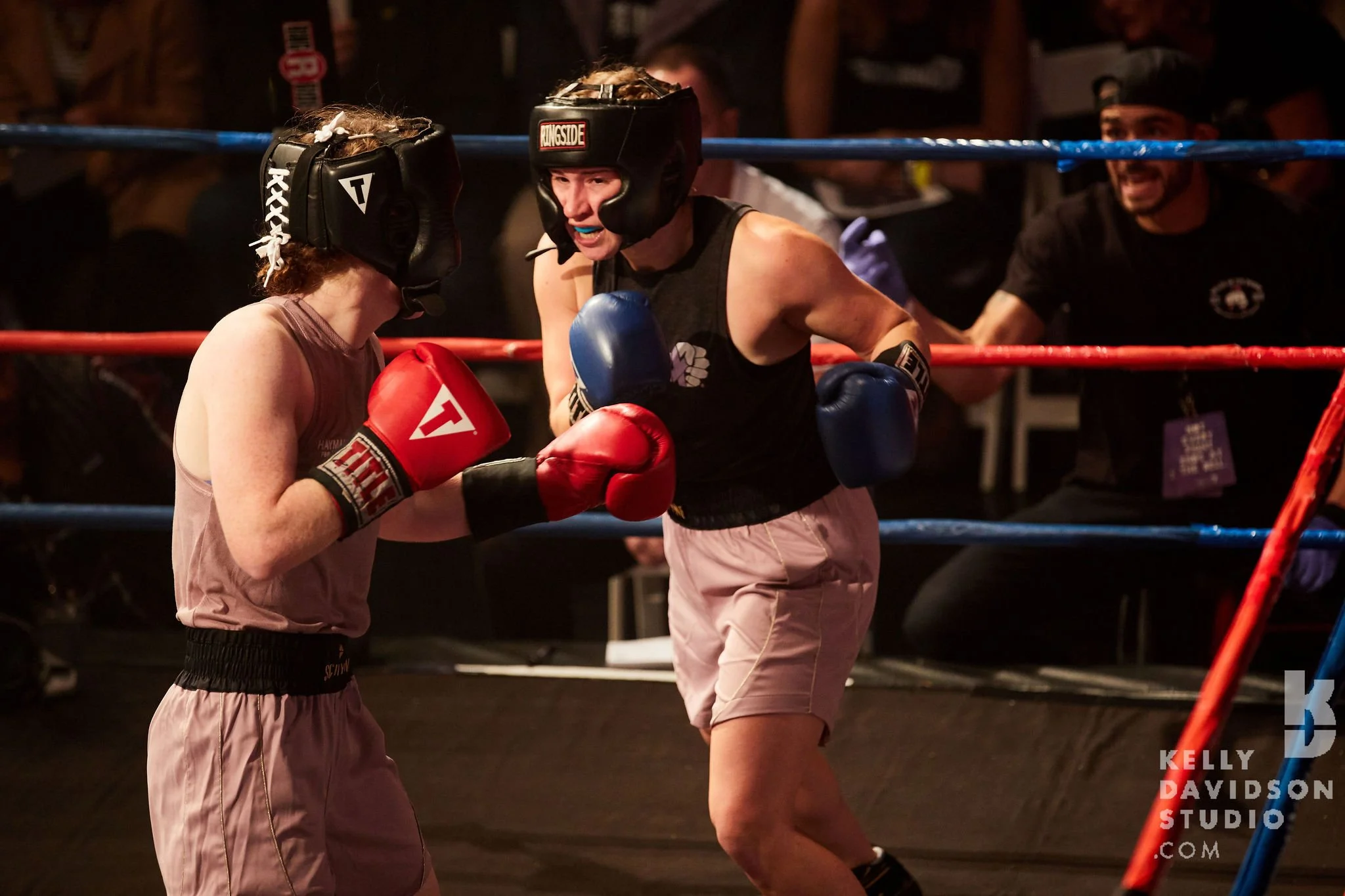 Two women boxers wearing protective gear sparring inside a boxing ring, with one woman throwing a punch at the other's gloves, while a referee observes in the background.