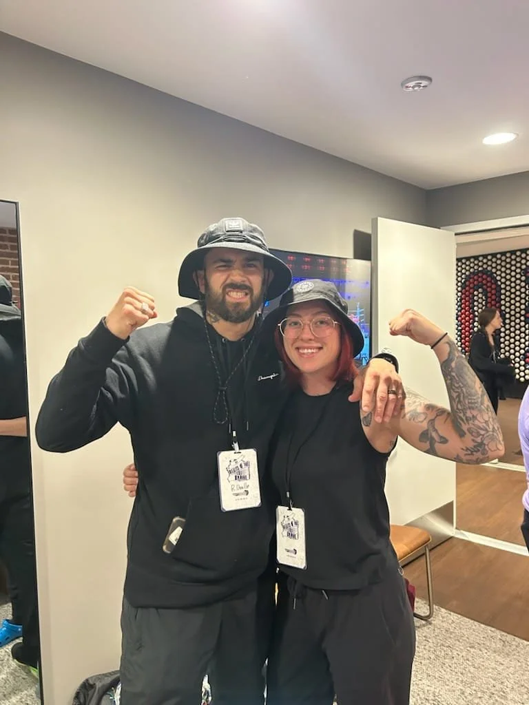 Two people, a man and a woman, posing together in a casual indoor setting, both flexing their arms and smiling. The man has a beard and is wearing a bucket hat, hoodie, and name badge. The woman has red hair, glasses, tattoos on her arm, and is also 