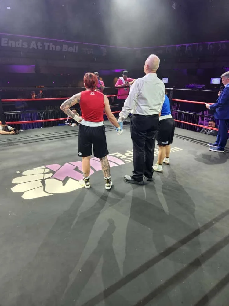 A female boxer with tattoos on her legs and arm, wearing a red sleeveless top and black shorts, stands in a boxing ring with her back to the camera, holding hands with an official. The ring has a logo on the floor, and there are two men in suits and 