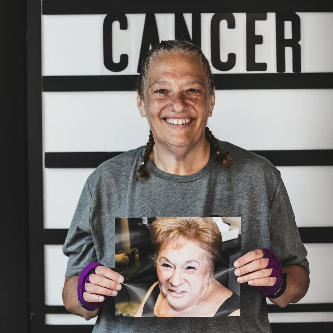 A smiling woman with braided hair holds a photo of an older woman with short, light-colored hair, in front of a black and white background with the word 'CANCER' partially visible.