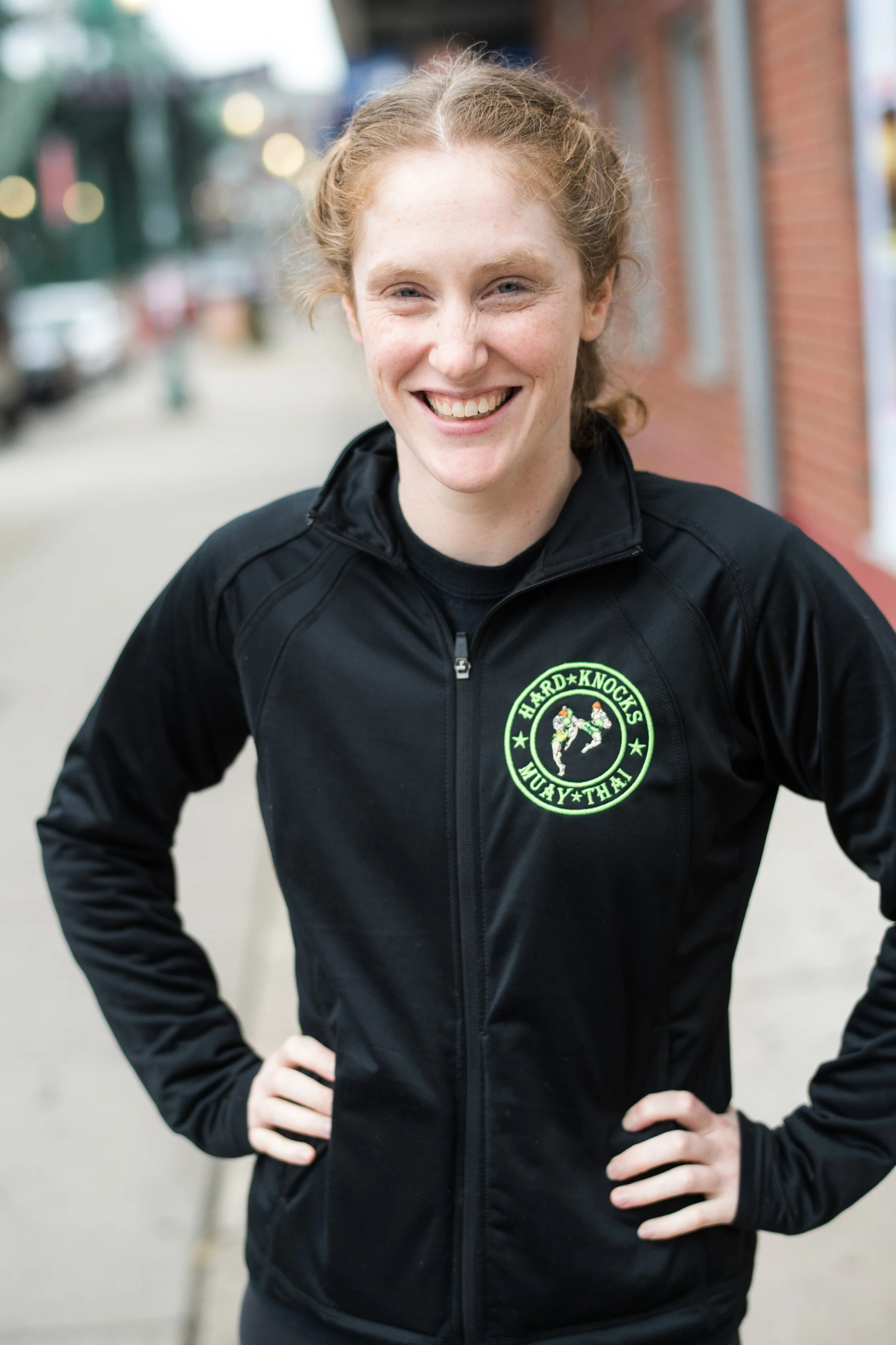 A young woman with red curly hair smiling outdoors, wearing a black jacket with a logo that says 'HARD KNOCKS MUAY-THAI' and features two fighters in a martial arts pose.