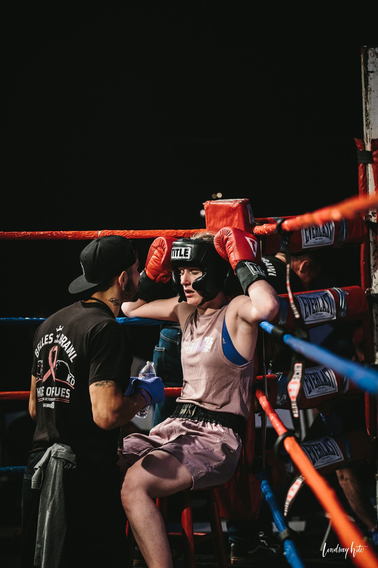 Female boxer sitting on a stool in her corner, wearing headgear and boxing gloves, talking to her coach during a boxing match.