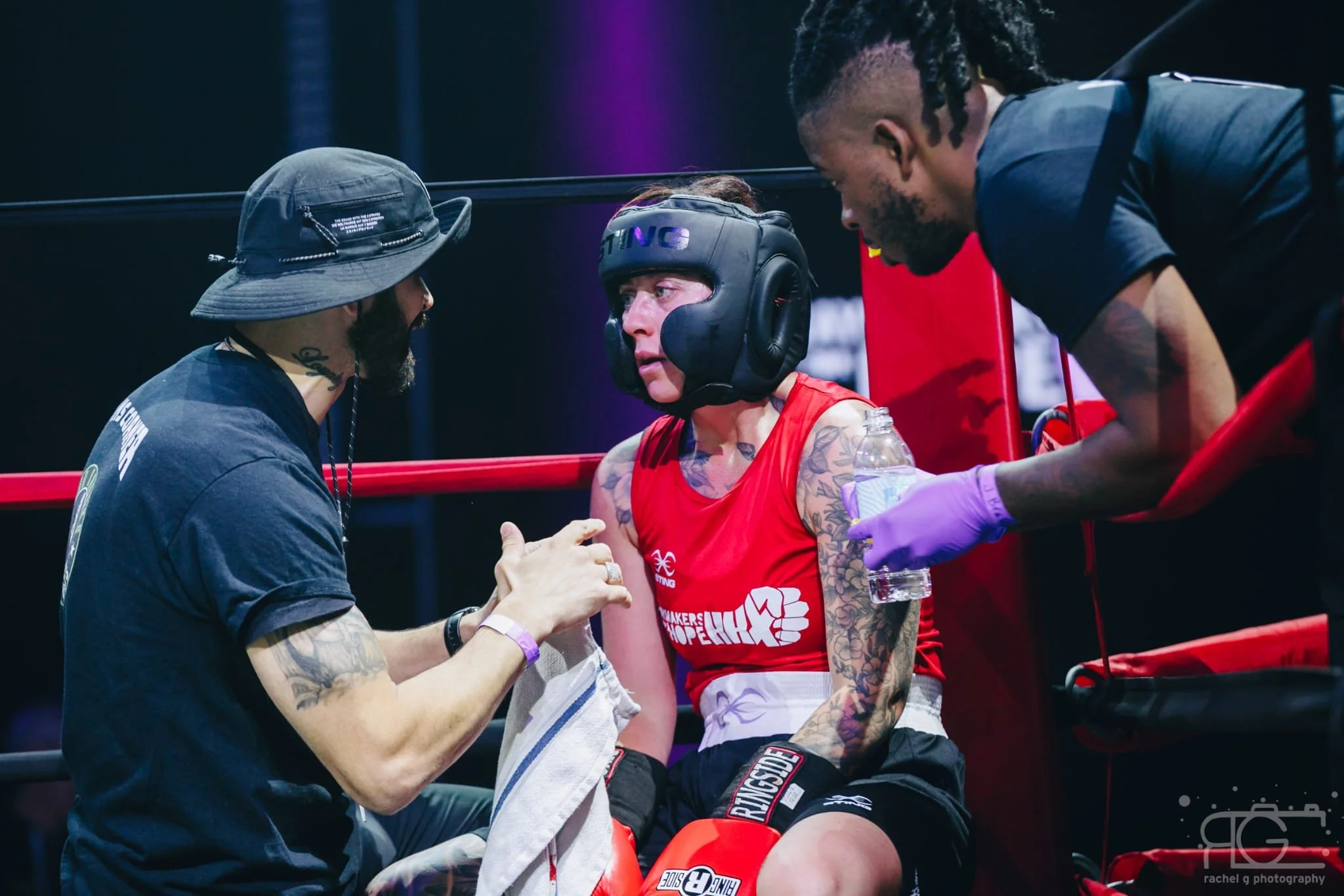 A female boxer in a red tank top and black shorts sits in the corner of a boxing ring, wearing a protective headgear, with her face showing signs of injury and exhaustion. Two trainers attend to her, one holding her hand and the other offering water,