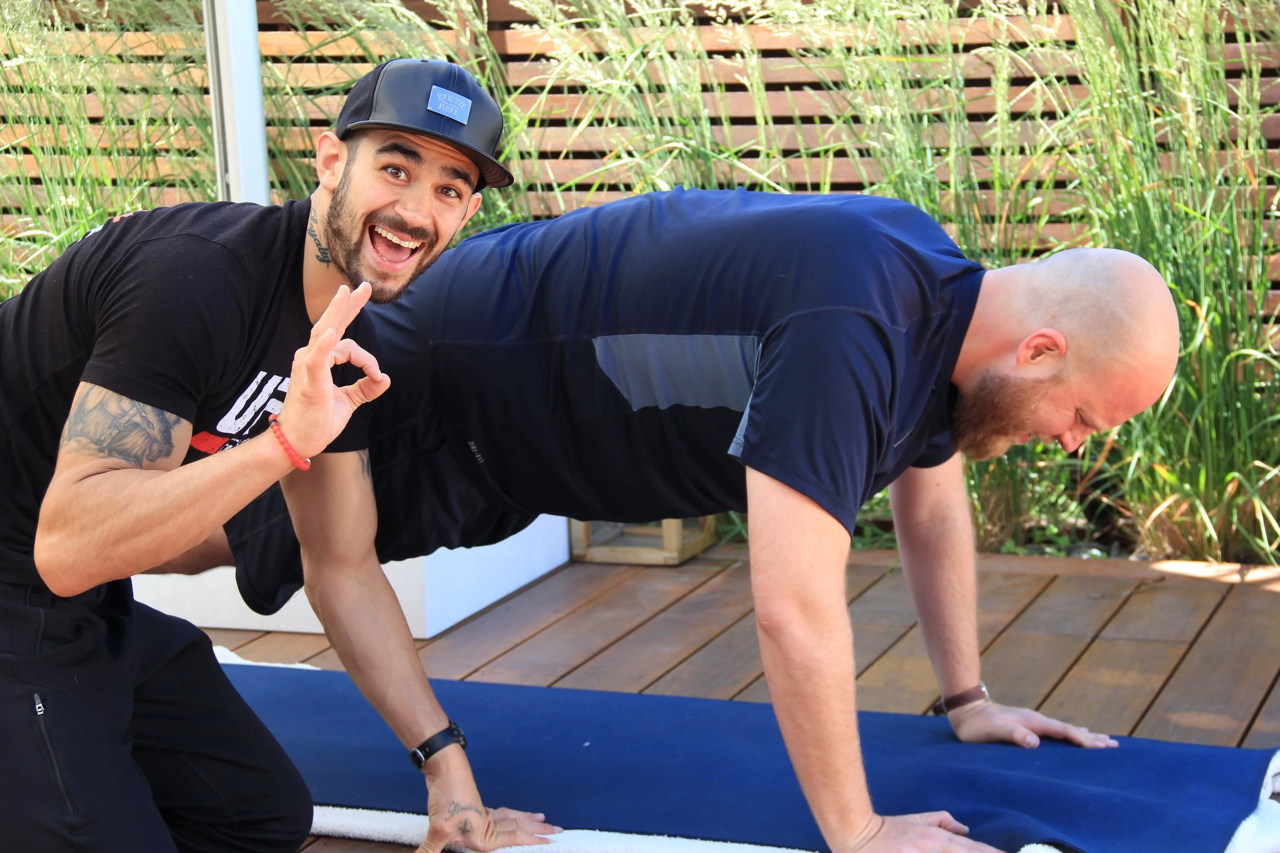 Two men practicing yoga outdoors on a wooden deck with greenery in the background. One man is in a plank position, and the other man is kneeling beside him, making an 'OK' hand gesture and smiling at the camera.