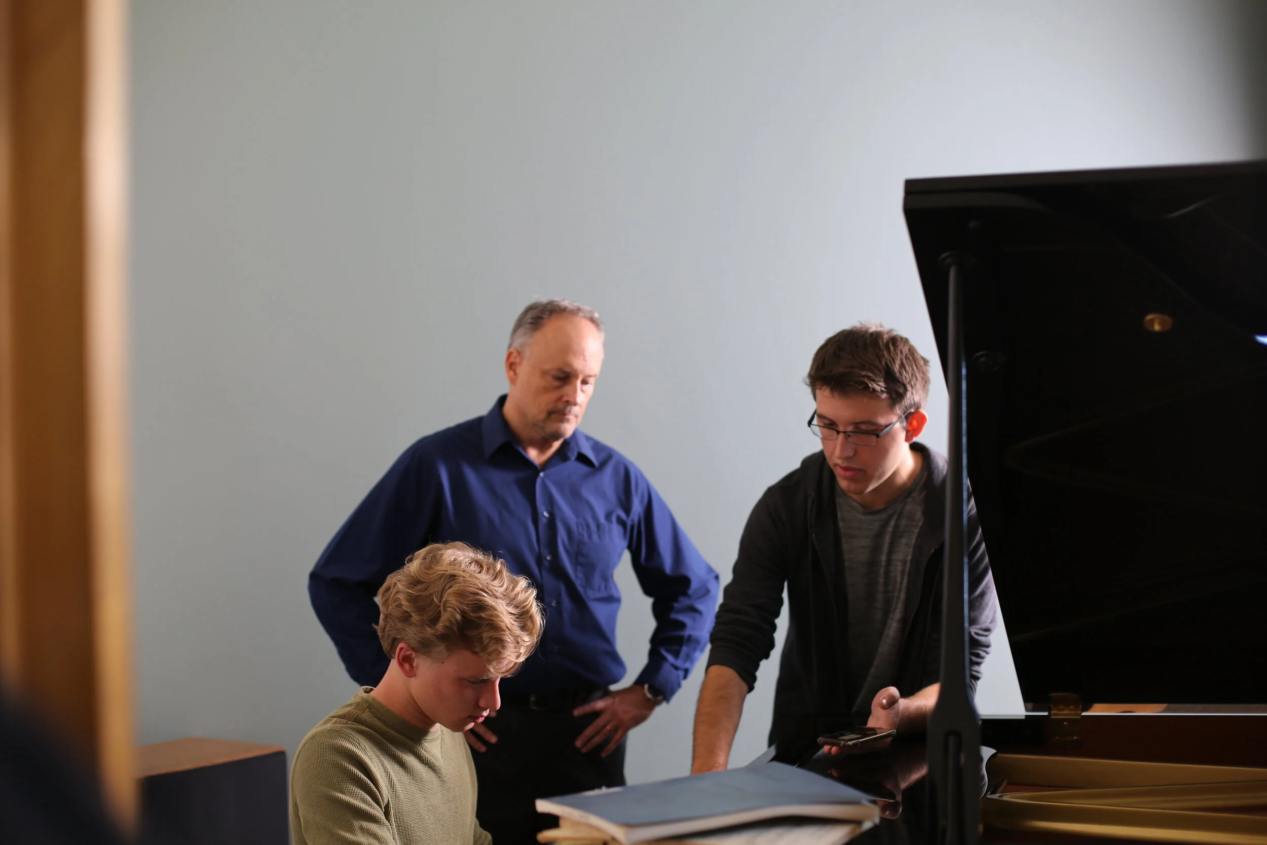 A young man with blonde hair playing a grand piano while three other males, two older and one younger, observe and discuss, in a room with plain walls.