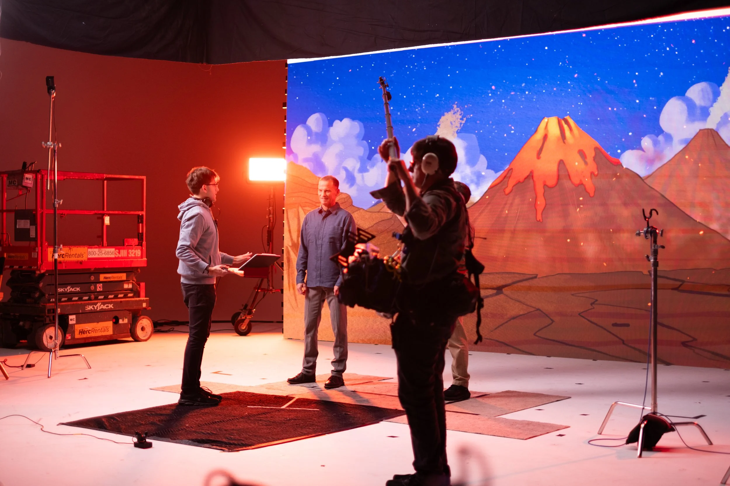 People on a film or television set with a large animated mountain background of erupting volcanoes and a starry sky, and equipment such as lights and scaffolding.