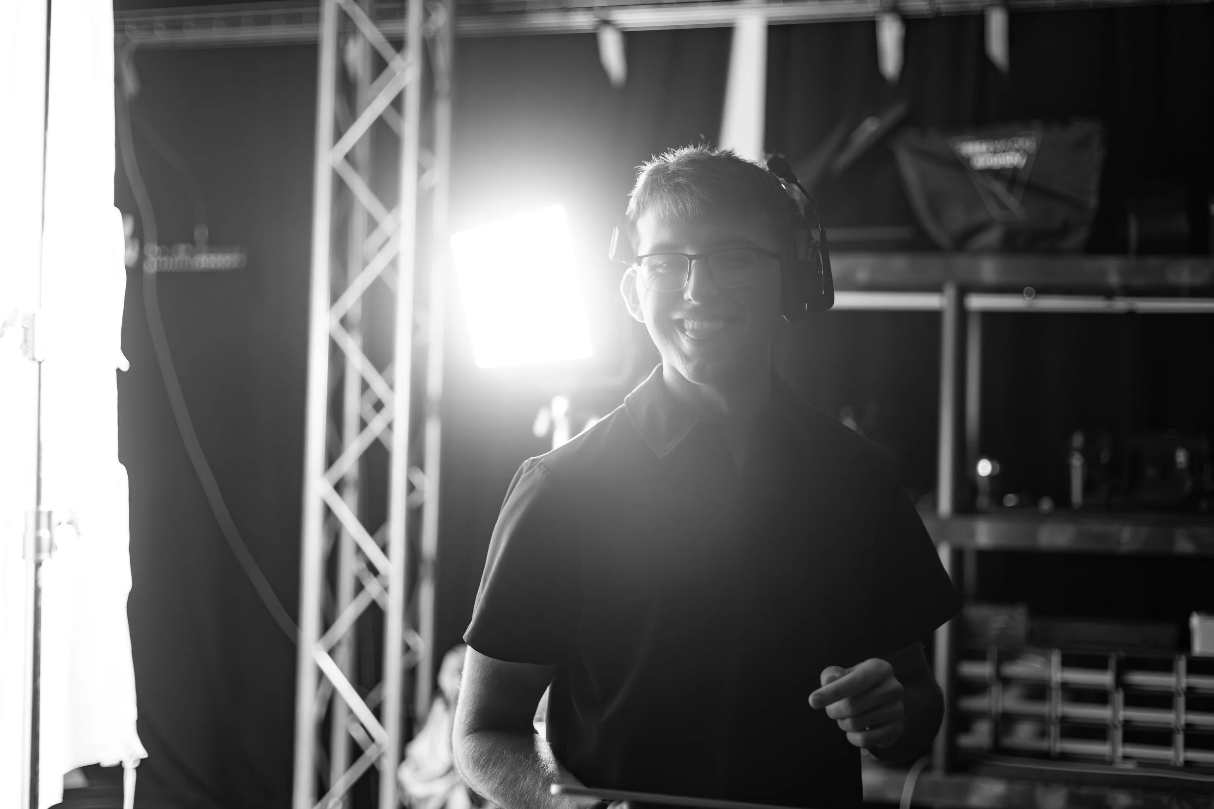 A young man with short hair, glasses, and headphones, smiling while standing in a backstage or technical area with shelving and metal frame structures, illuminated by bright backlighting.